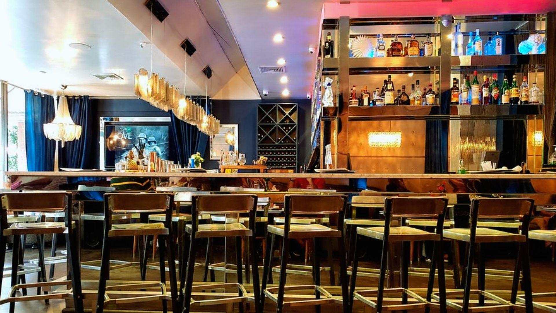 Modern upscale bar with mirrored counter, wood stools, lit liquor shelves.
