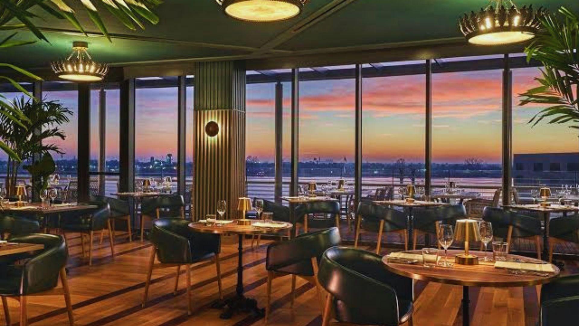 Upscale rooftop restaurant with wooden tables, green chairs and floor-to-ceiling windows overlooking sunset over the river.
