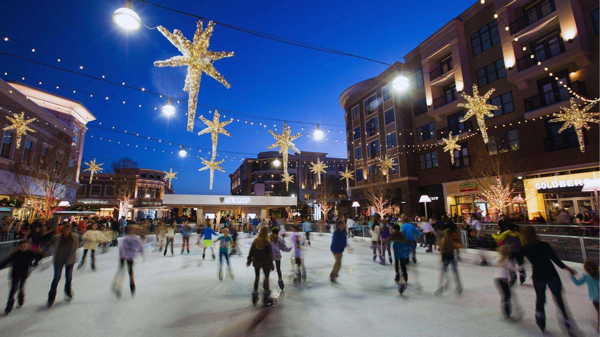 Ice skating rink at dusk with glowing star lights overhead and crowd skating near shops.