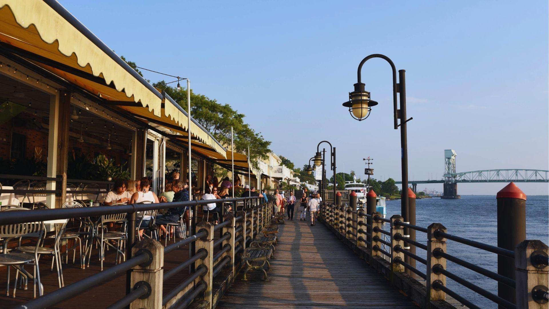 Restaurant patio with diners under yellow awnings along wooden boardwalk overlooking river and bridge view.