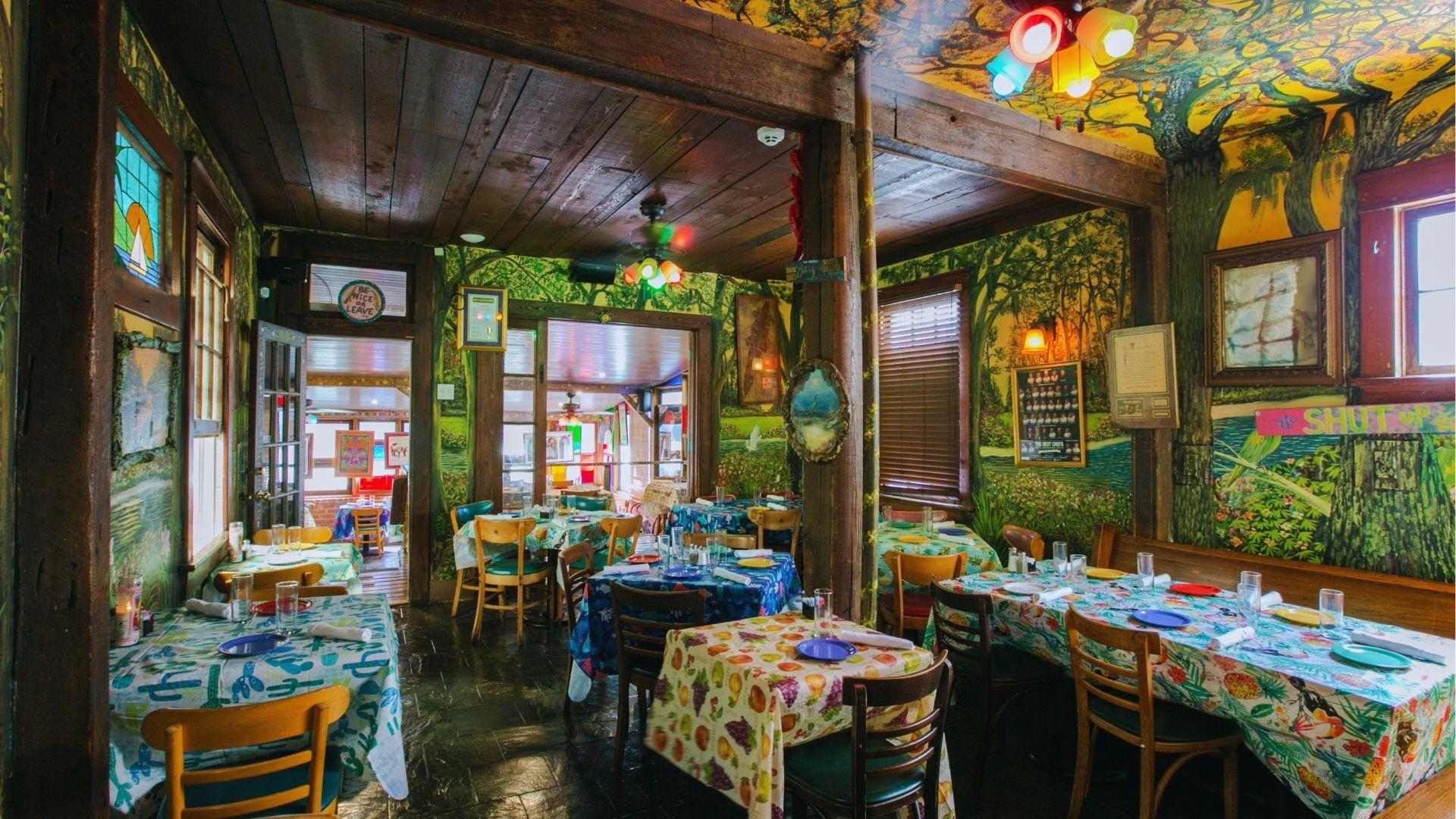 Eclectic colorful restaurant with forest murals, fruity tablecloths, wooden chairs and vibrant stained glass decor.