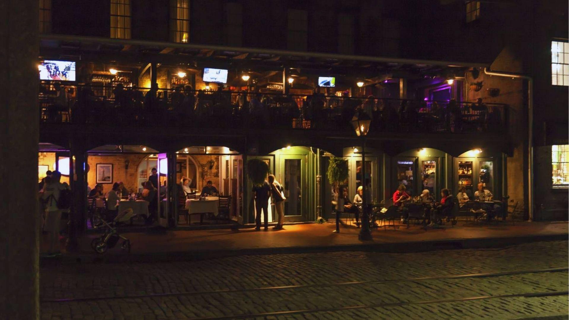 Lively nighttime outdoor dining scene on cobblestone street with illuminated restaurant balconies and patrons gathering.