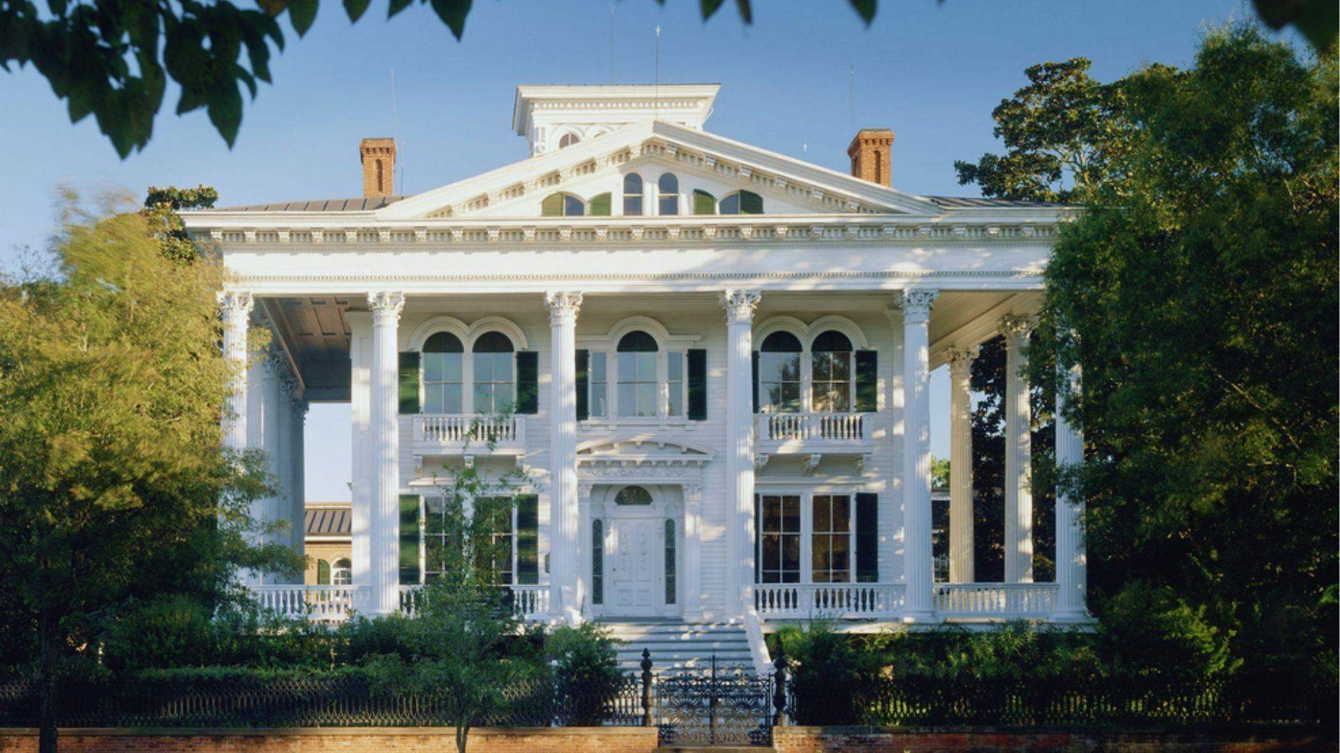 White antebellum mansion with tall Corinthian columns, green shutters and grand staircase surrounded by trees.