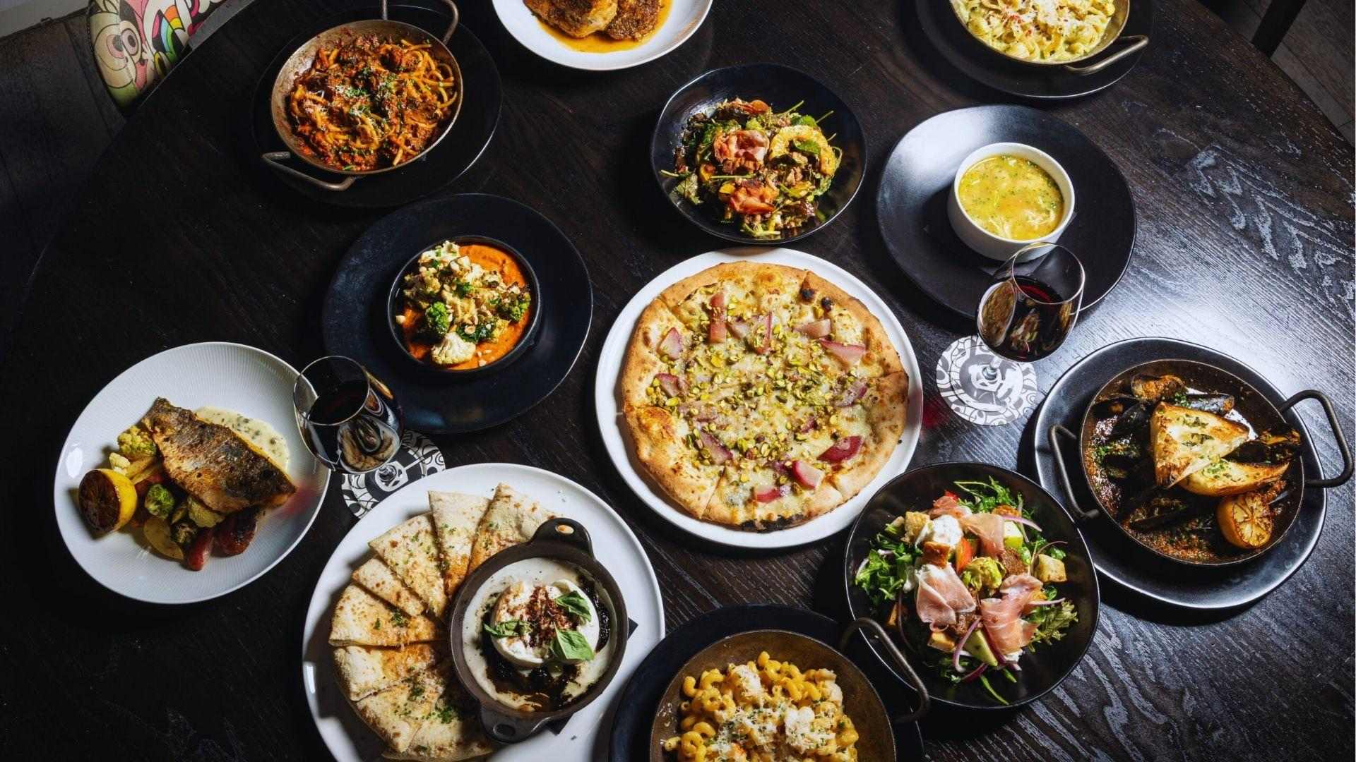Top-down spread of gourmet pizza pasta salads seafood dishes served on dark wooden restaurant table.