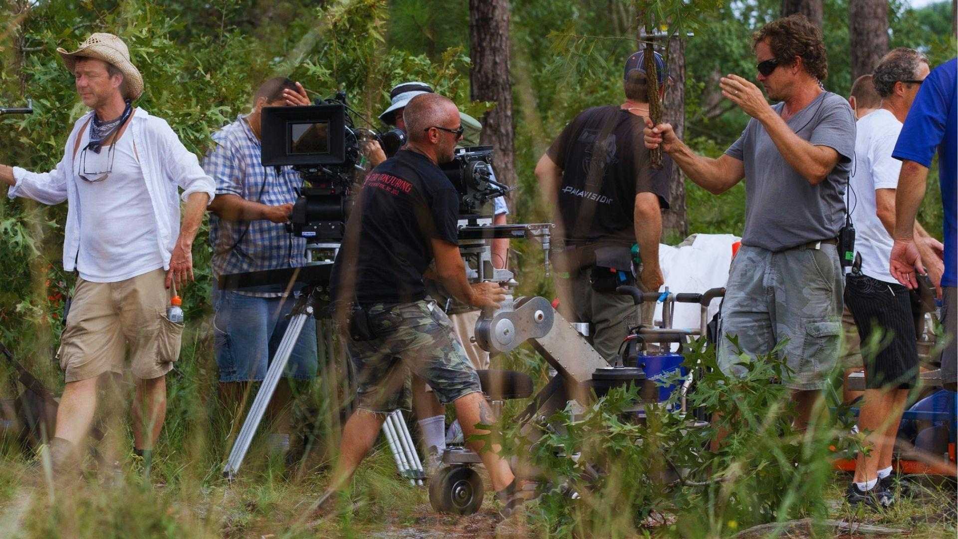 Film crew operating large camera on dolly in wooded area during outdoor movie production shoot.