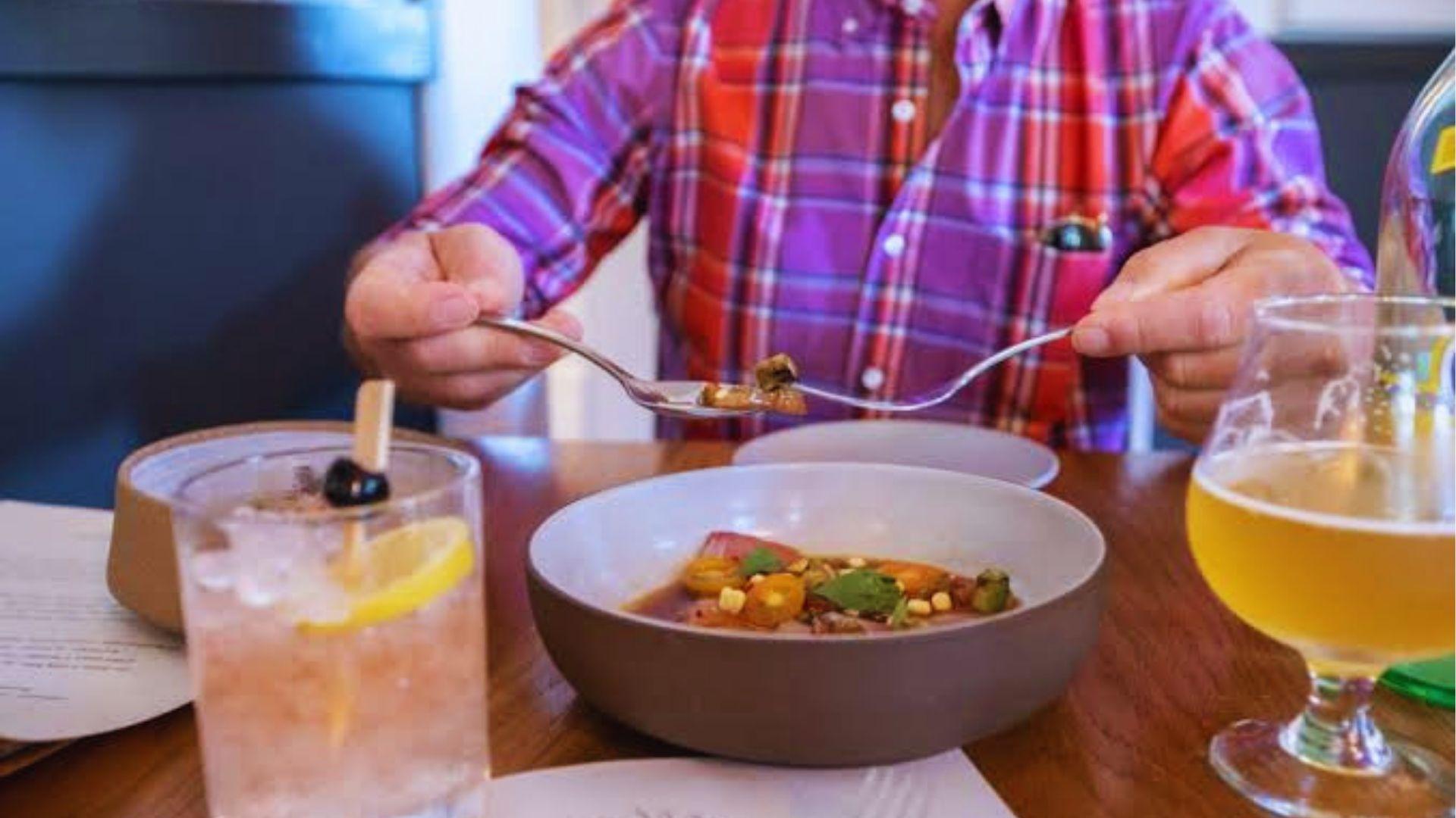 Person in plaid shirt sharing colorful dish with drinks on wooden restaurant table indoors today.