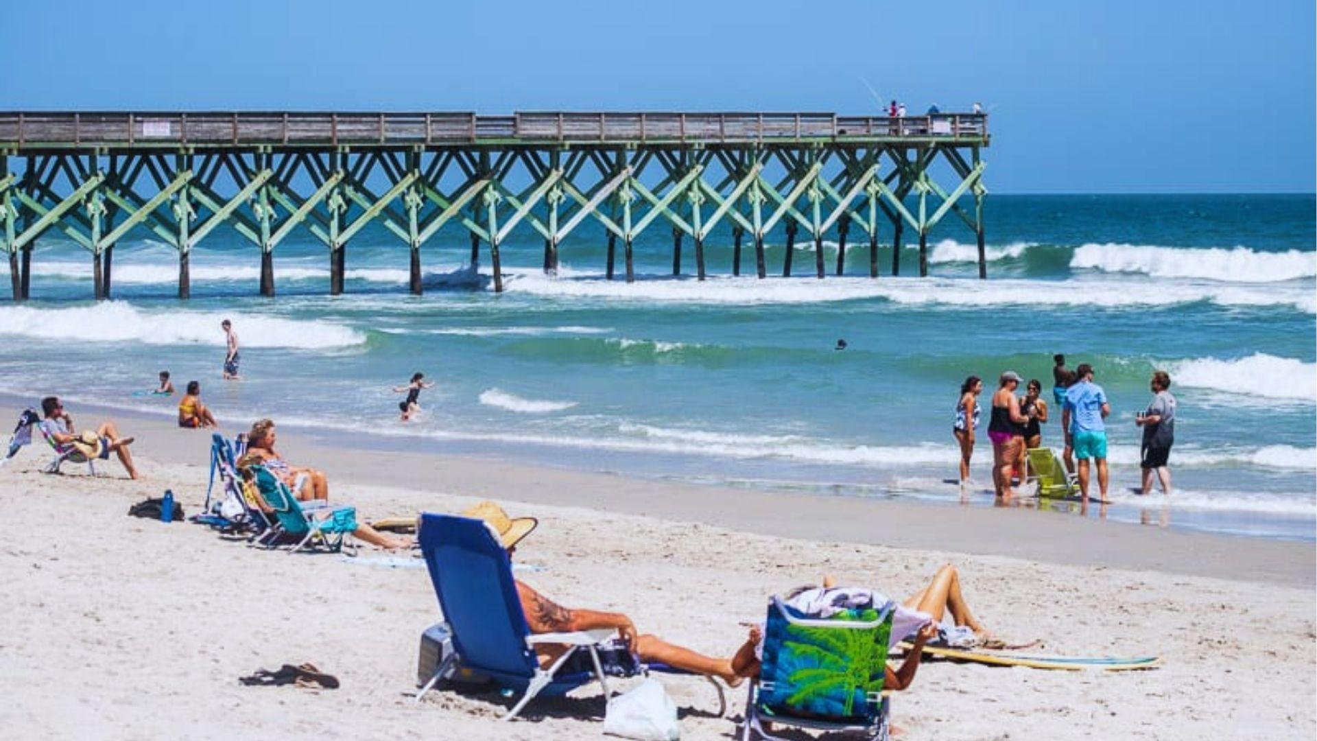 Beachgoers relaxing in colorful chairs on a sandy shore near a fishing pier with waves rolling in.