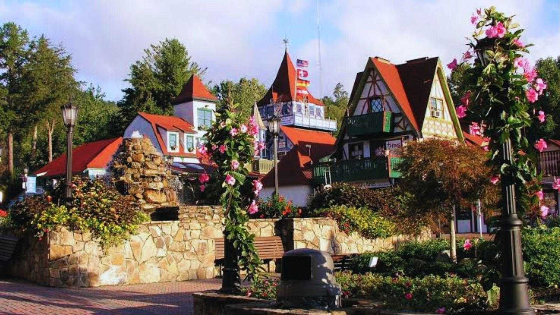 Bavarian village square with stone fountain, pink flowering vines, red-roofed buildings and lush green trees.