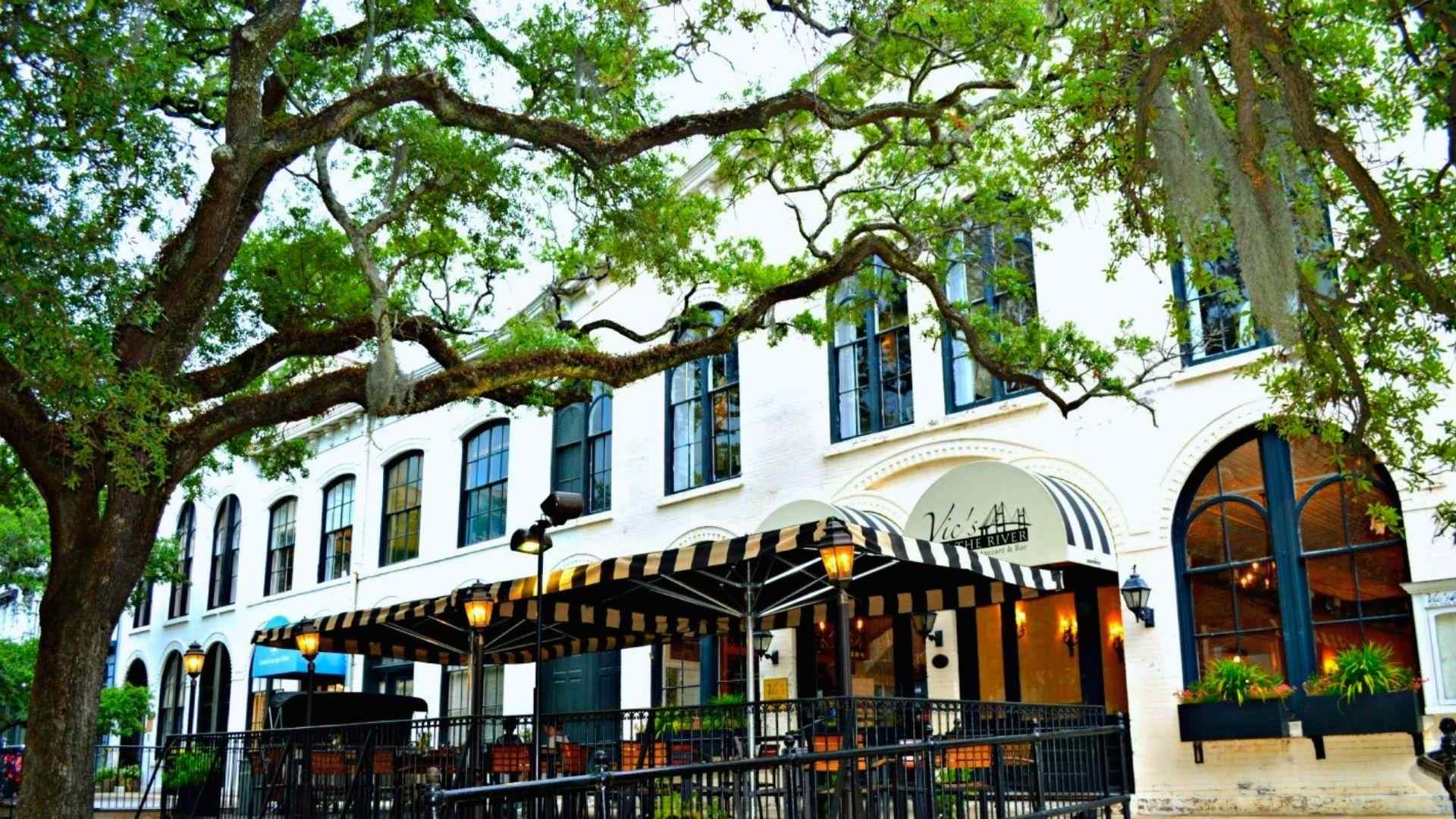 White historic riverfront restaurant Vic's On The River with striped awnings, outdoor patio, large oak tree with Spanish moss.