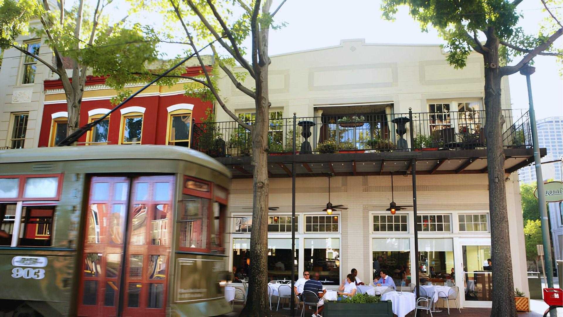 Green streetcar numbered 903 passing in front of an outdoor restaurant with balcony diners and trees.