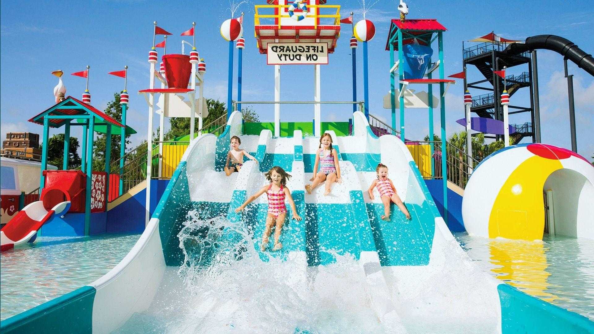 Four children sliding down bright water park slides with colorful lifeguard tower and splash buckets
