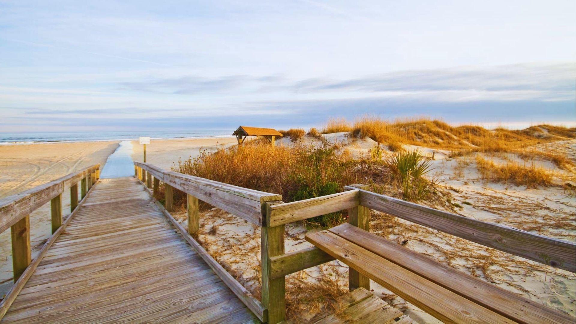 Wooden boardwalk with bench leading over dunes to sandy beach under cloudy sky today peacefully.