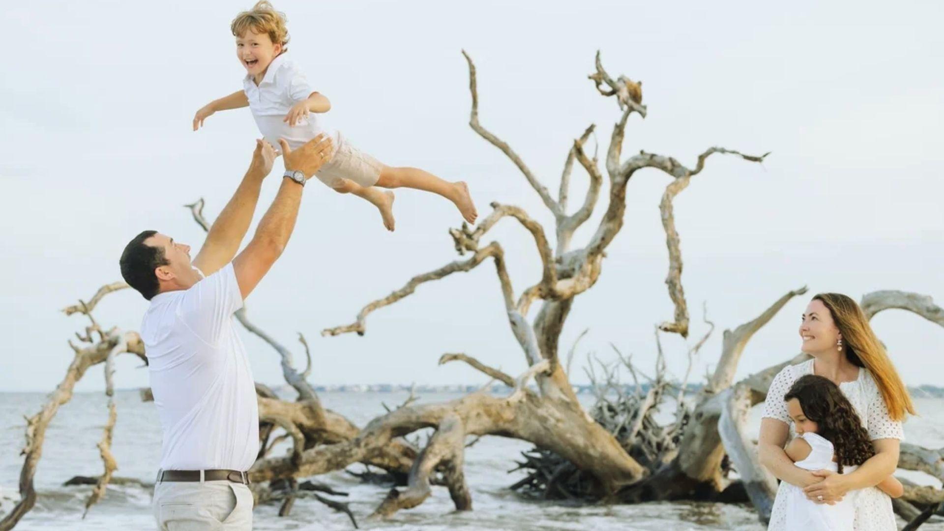 Family of four in white on beach with driftwood, father lifting smiling boy in air, mother hugging young girl.