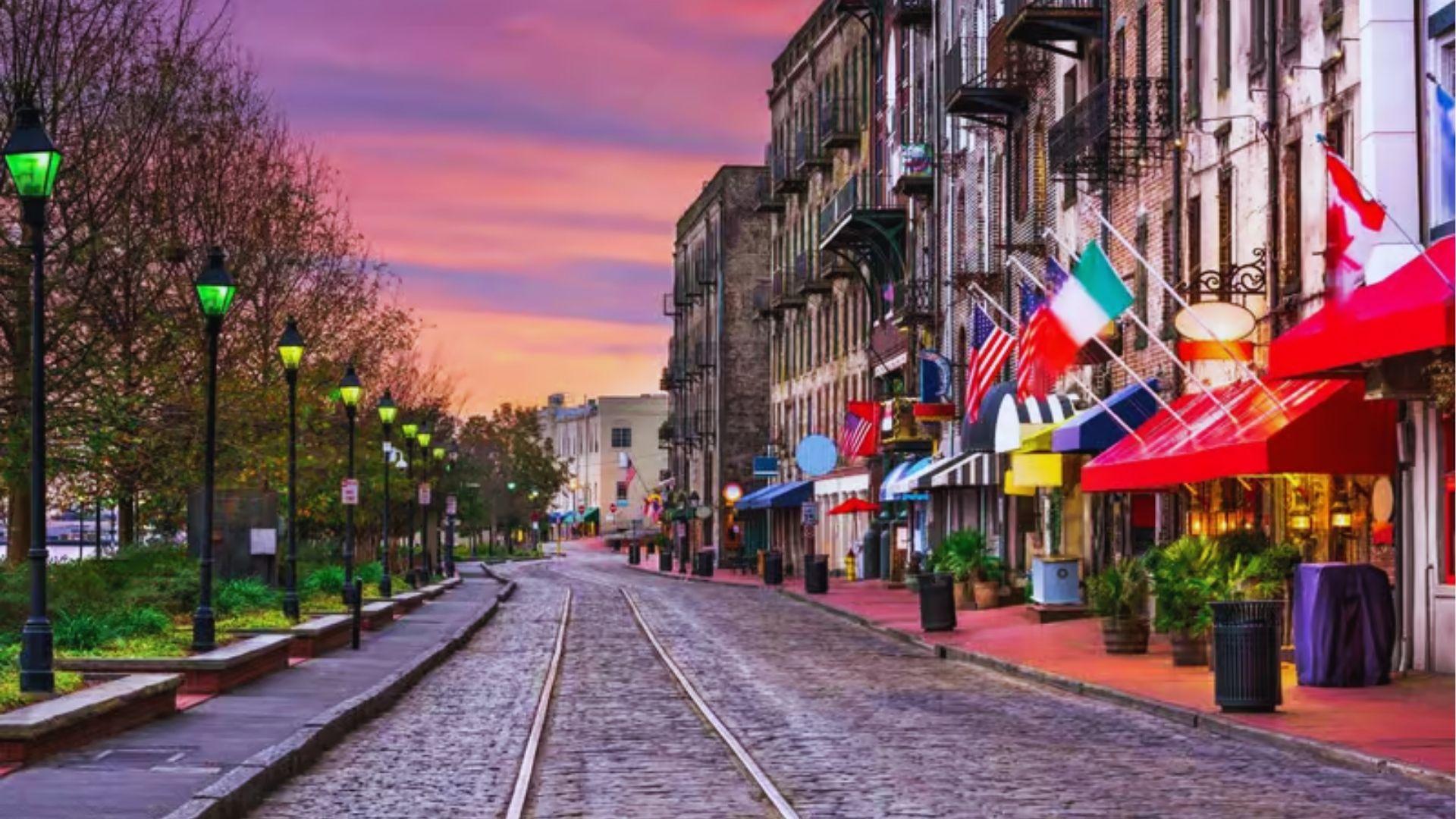Cobblestone River Street at sunset with colorful awnings, flags, lampposts, and historic brick storefronts glowing.