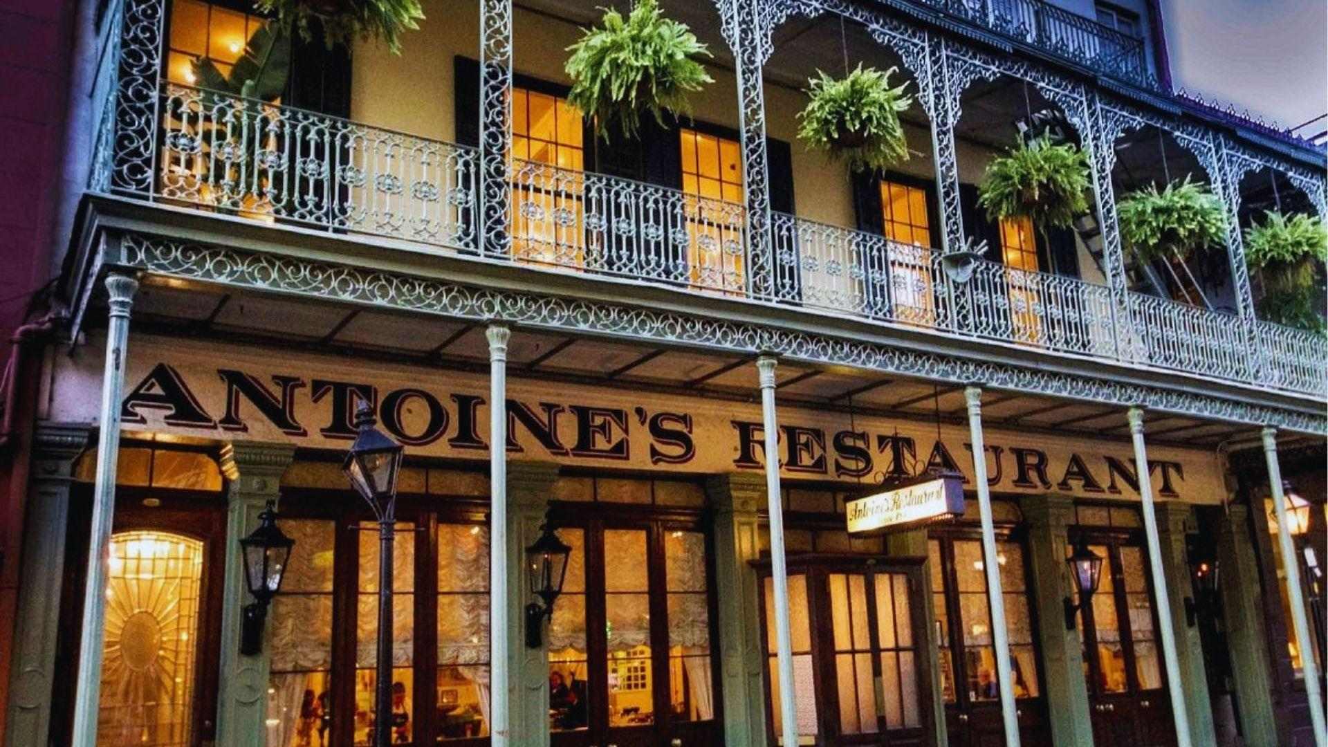 Historic Antoine's Restaurant facade with ornate iron balconies hanging ferns and glowing evening lights outside.