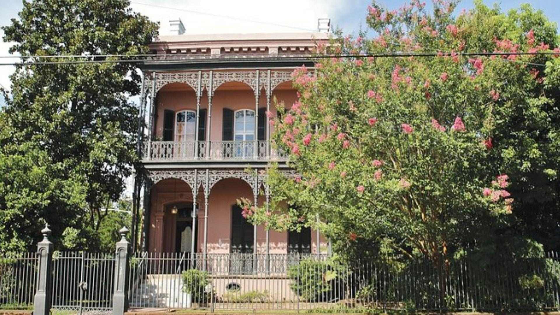 Historic pink double-gallery home with ornate iron balconies behind gated fence