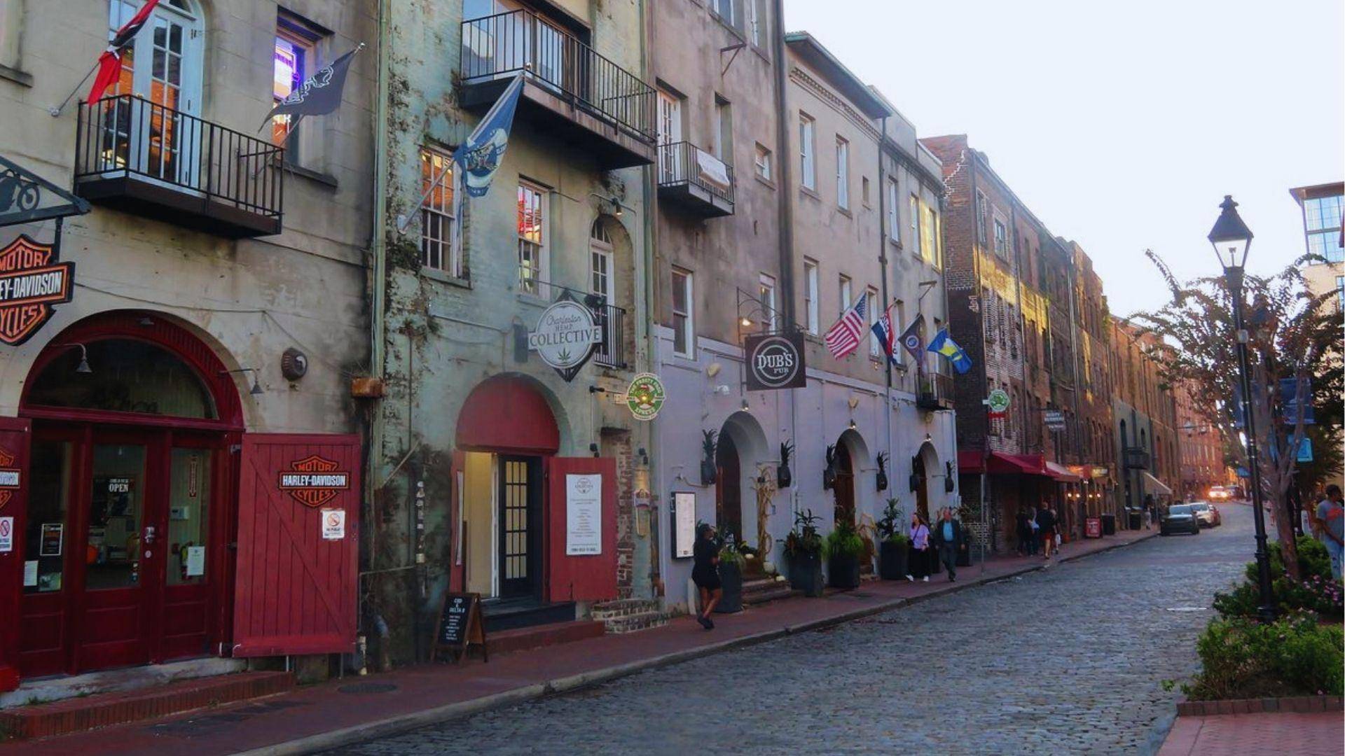 Cobblestone River Street lined with rustic shops, Harley-Davidson store and flags under evening sky today.