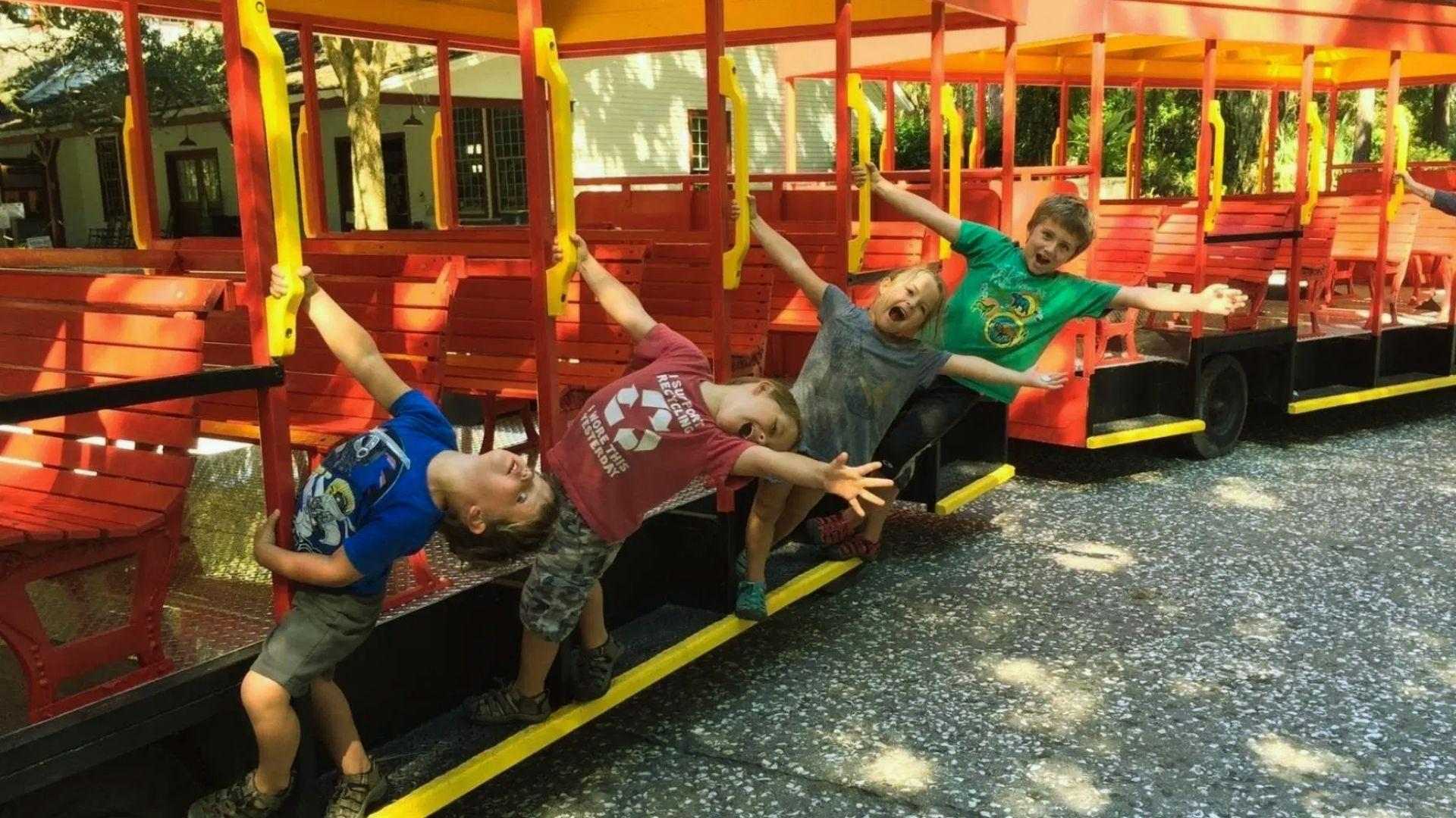 Four children joyfully leaning out from bright red open-air trolley car on sunny day