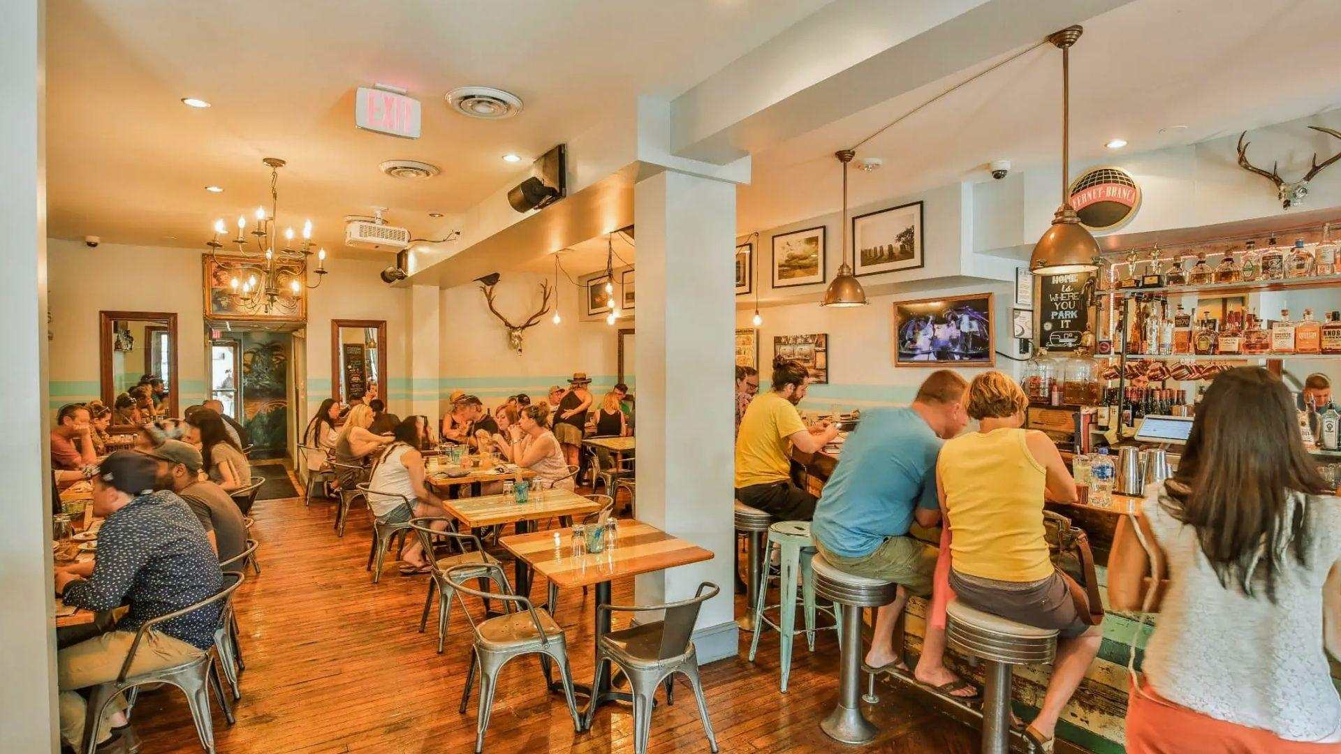 Busy rustic bar and dining room with patrons at tables and bar stools, wood floors, antler decor.
