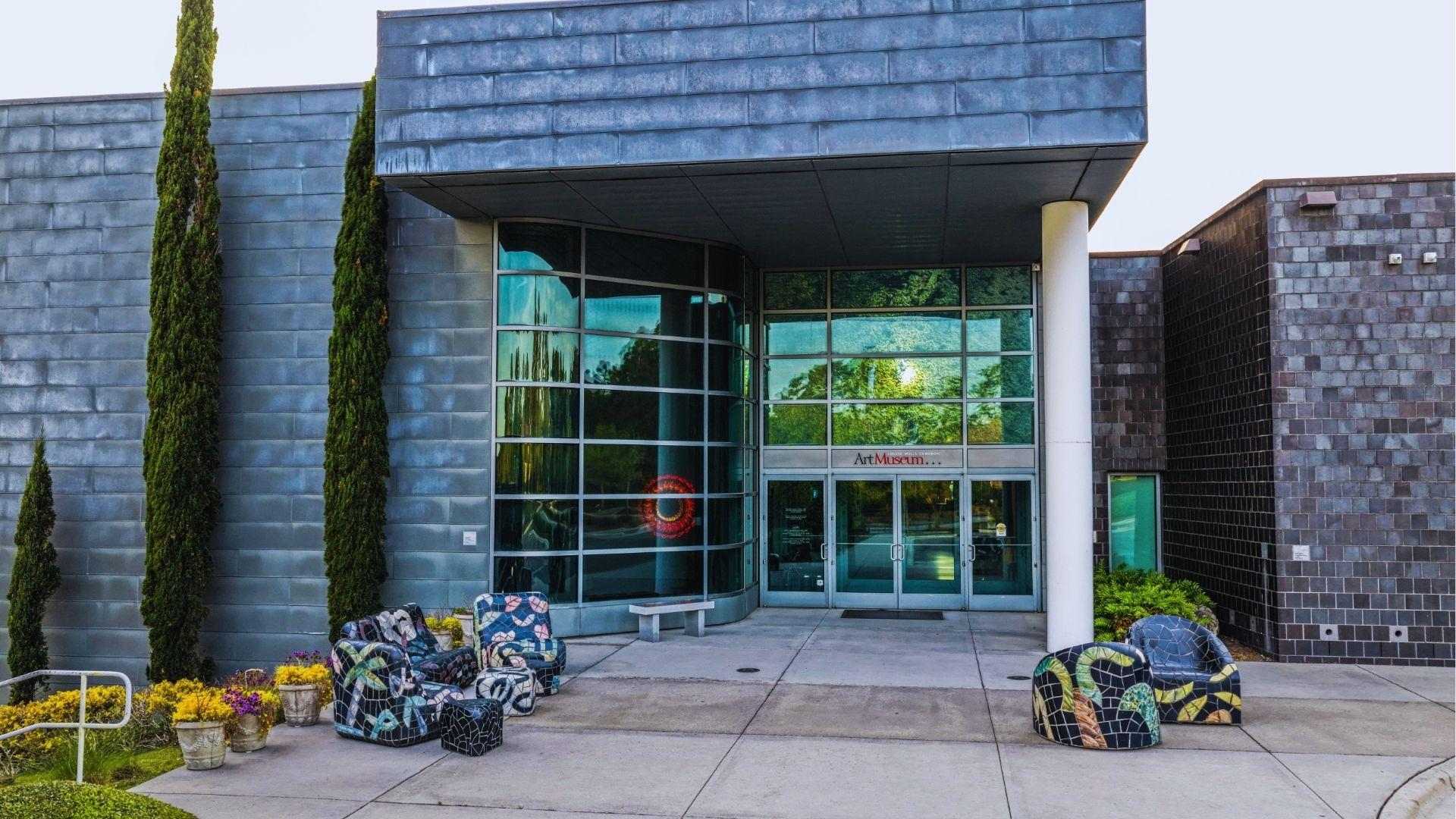 Art museum entrance with glass doors, mosaic chairs tall cypress trees and yellow potted flowers.