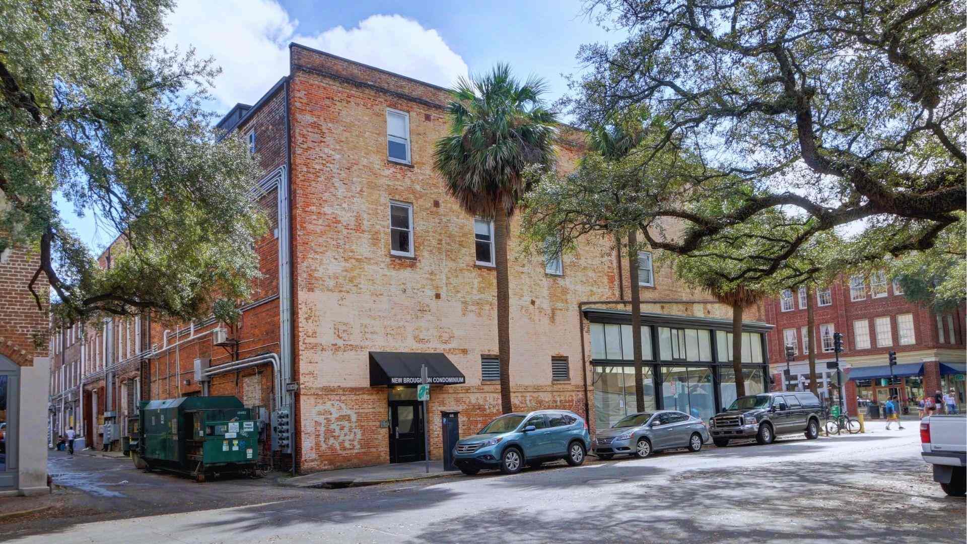 Weathered brick building housing New Broughton Condominium with palm trees and cars parked along the street.
