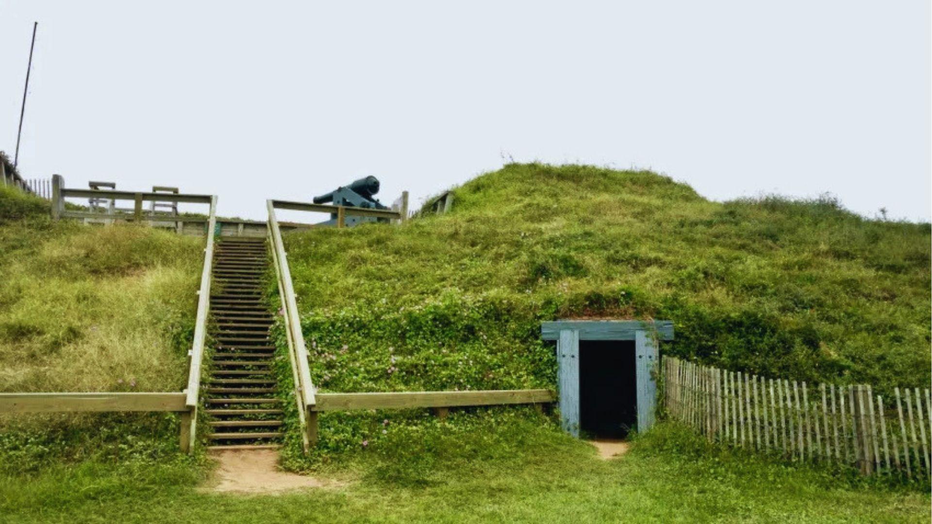 Historic grass-covered fort mound with wooden stairs, cannon on top and small entrance bunker below.