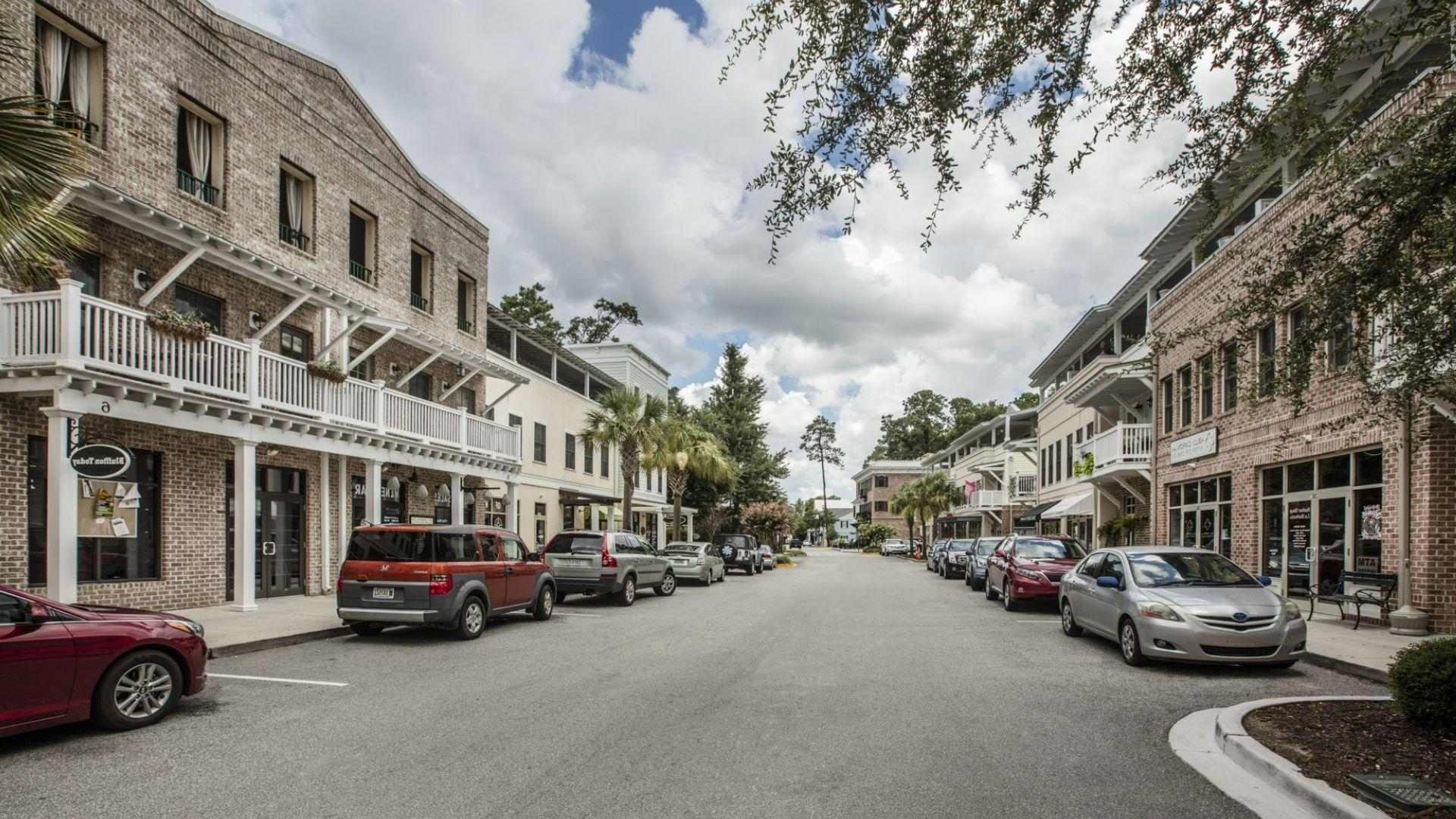 Charming brick main street with white balconies, parked cars, shops, palm trees under cloudy sky