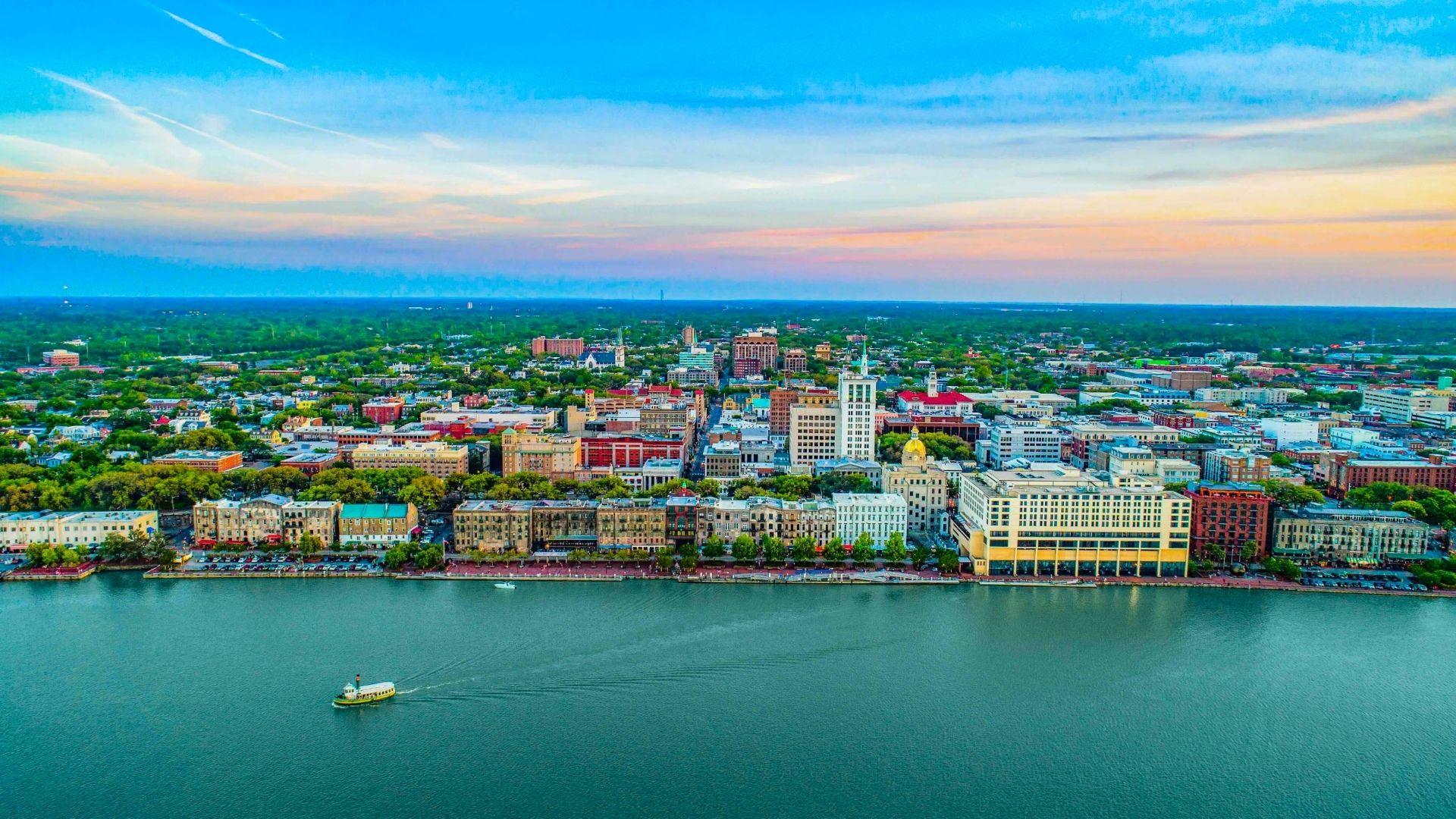 Aerial view of Savannah riverfront at sunset with boat on water and colorful downtown buildings