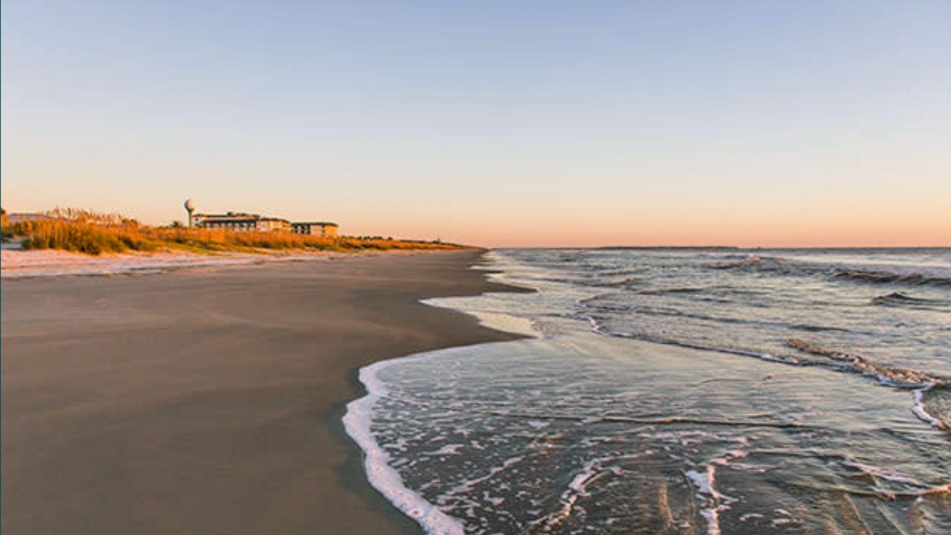 Gentle waves washing onto sandy beach at sunset with dunes and distant building