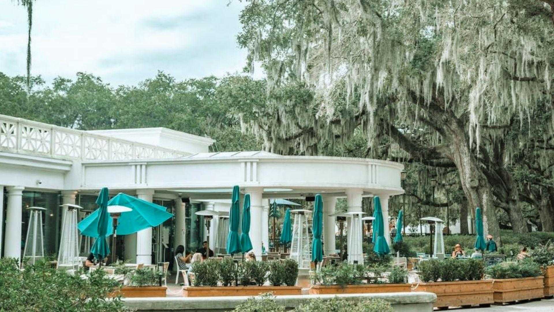 Outdoor restaurant patio with teal umbrellas, white colonnade, planters, and moss-draped oak trees.