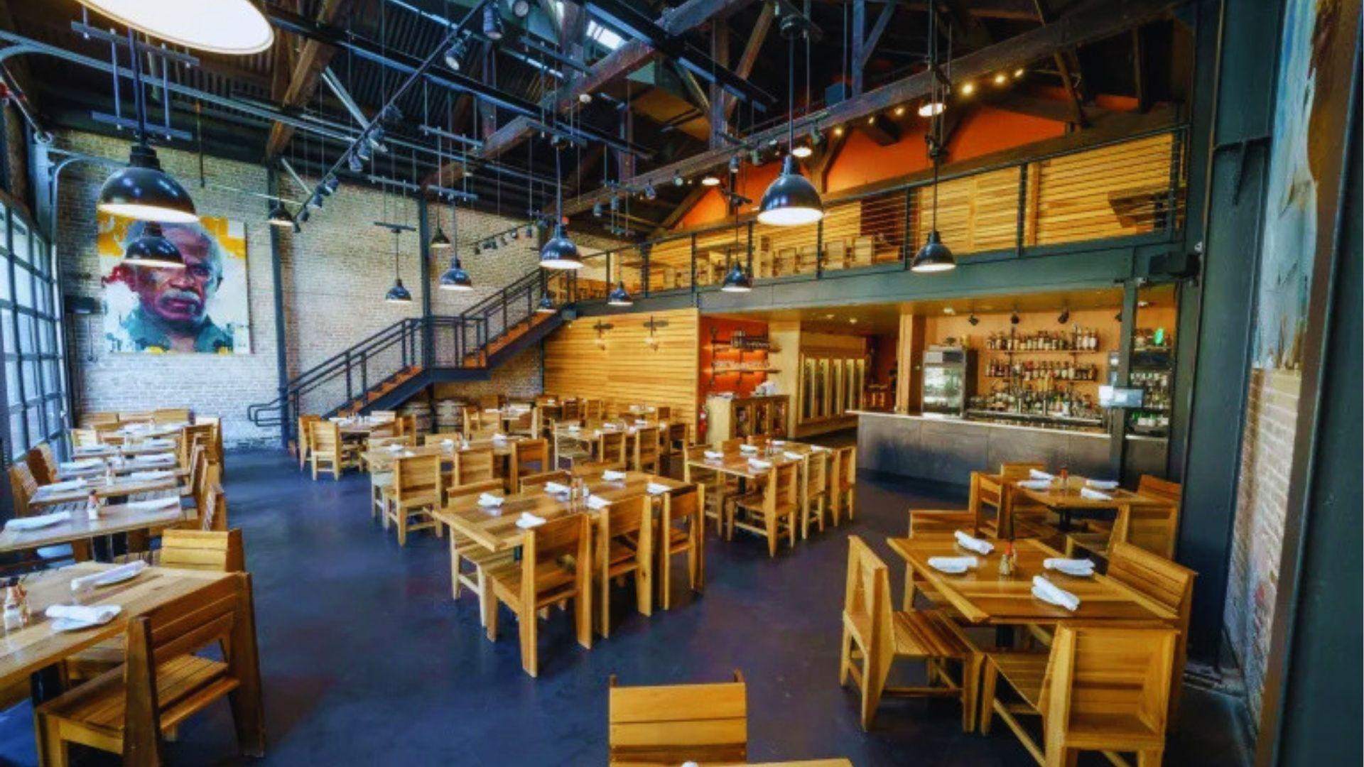 Spacious industrial restaurant with wooden tables black pendant lights, exposed beams and staircase to loft.