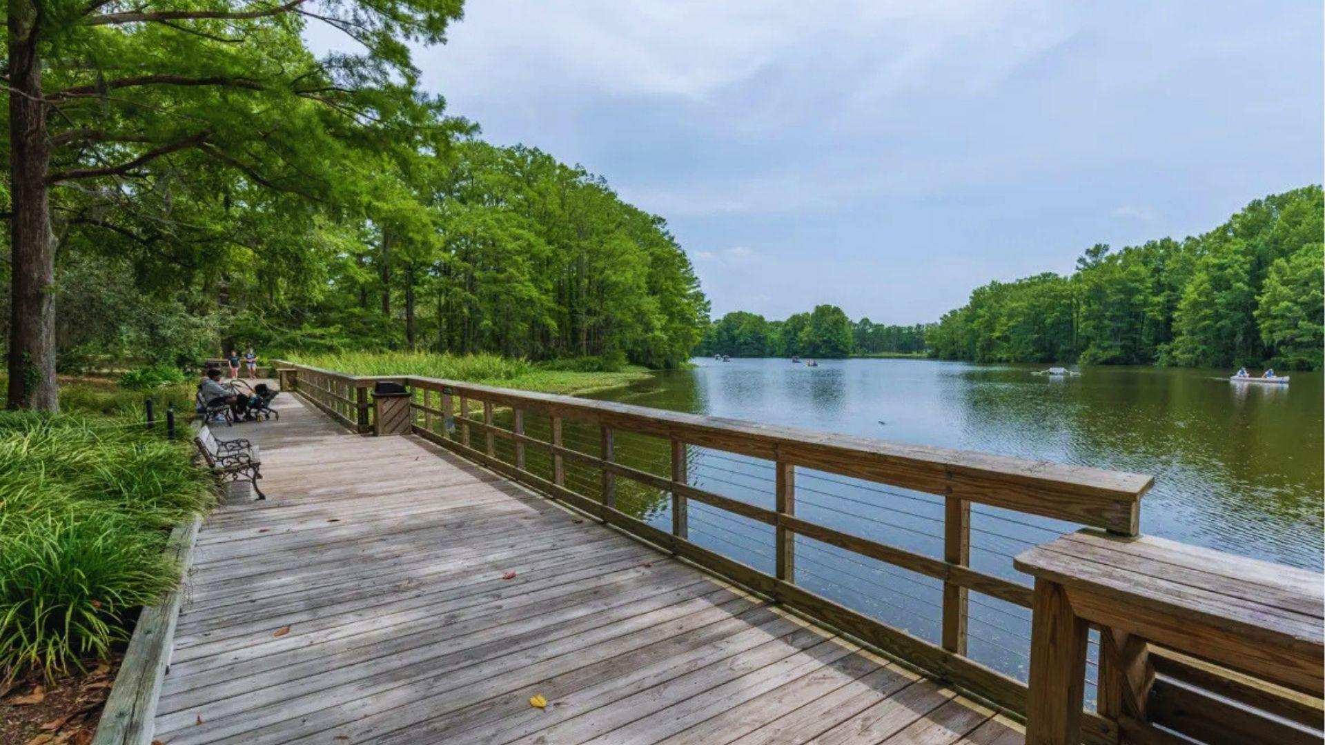 Wooden boardwalk stretching along serene lake surrounded by lush green trees with kayakers in distance.