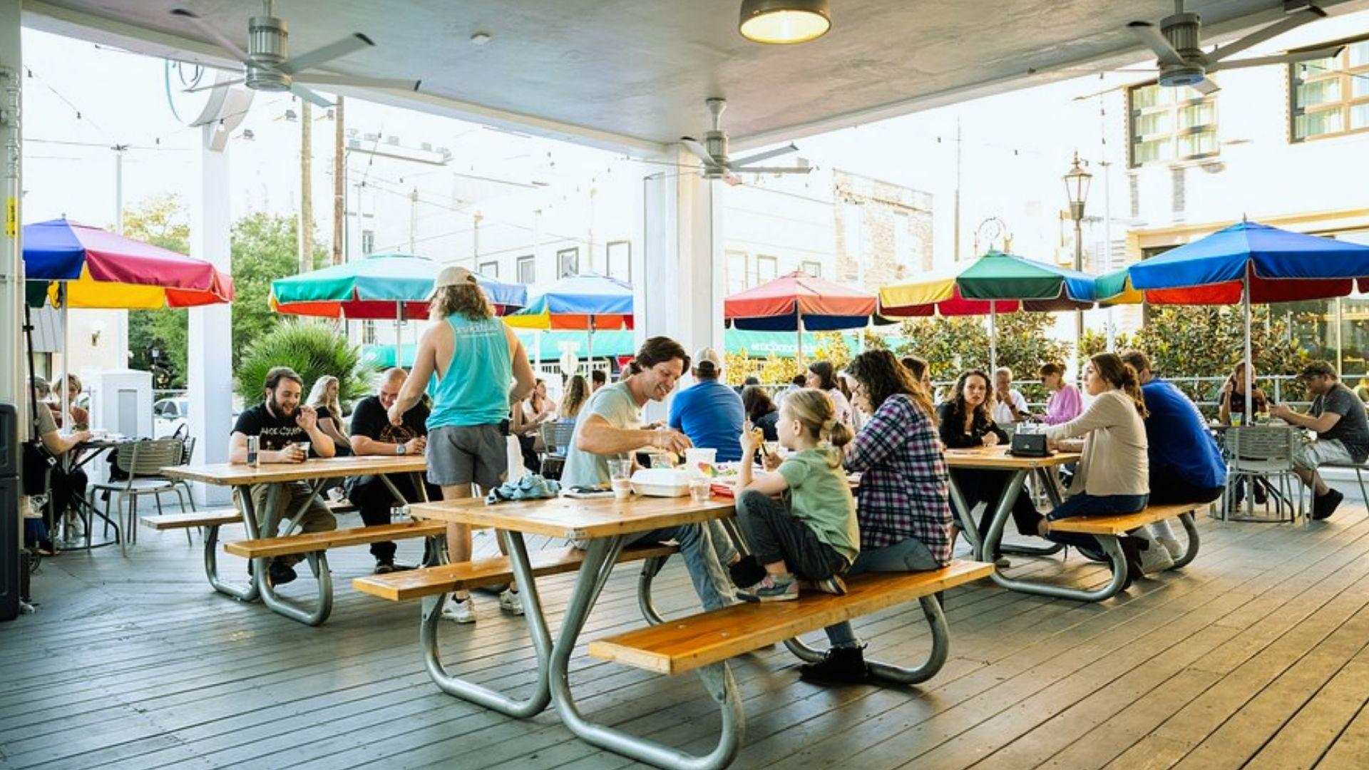 Covered outdoor patio with wooden picnic tables, families dining under colorful rainbow umbrellas, ceiling fans overhead.
