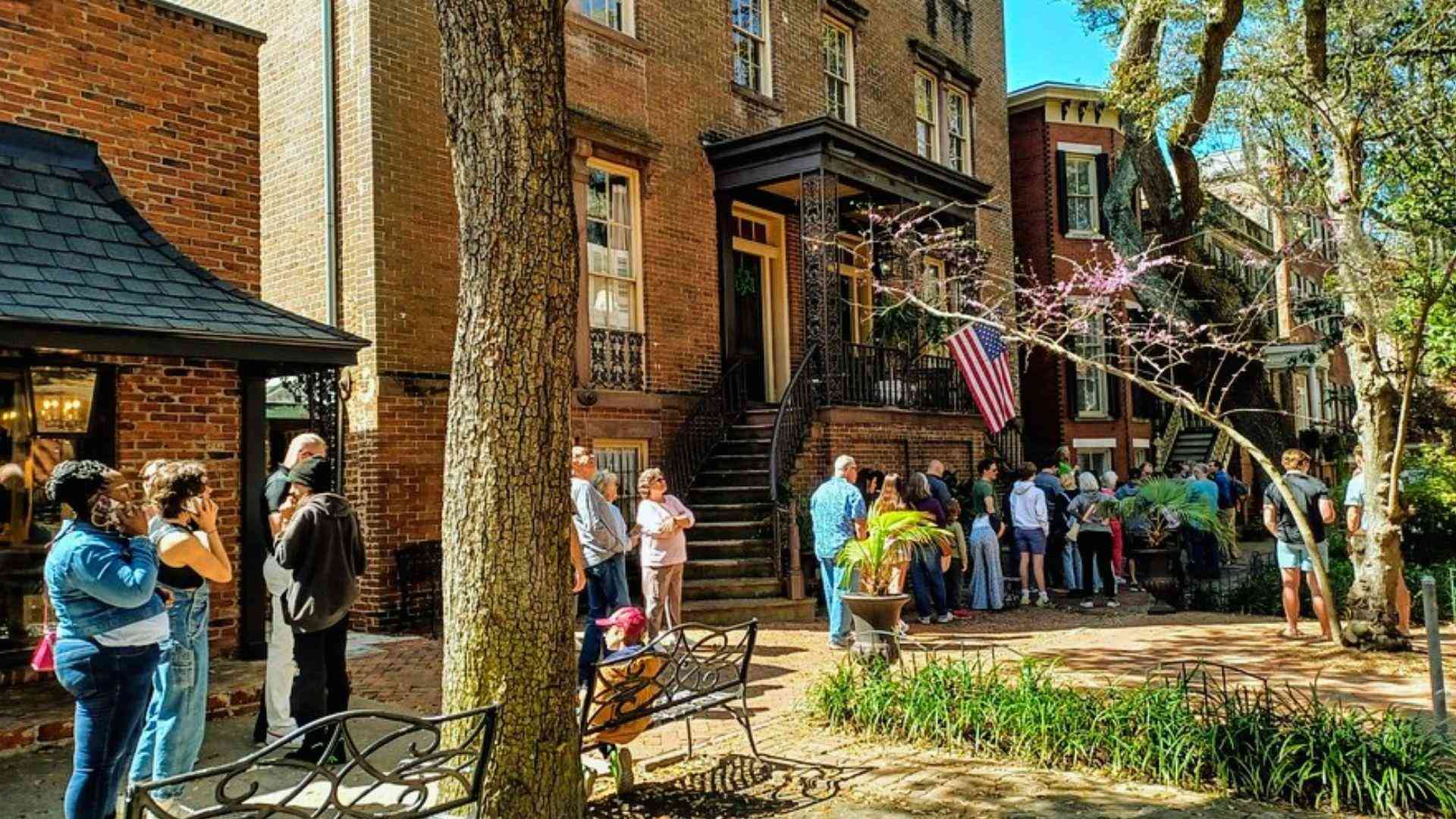 Sunny historic Savannah brick townhouses with tourists lined up outside, American flag, blooming redbud tree, iron benches.