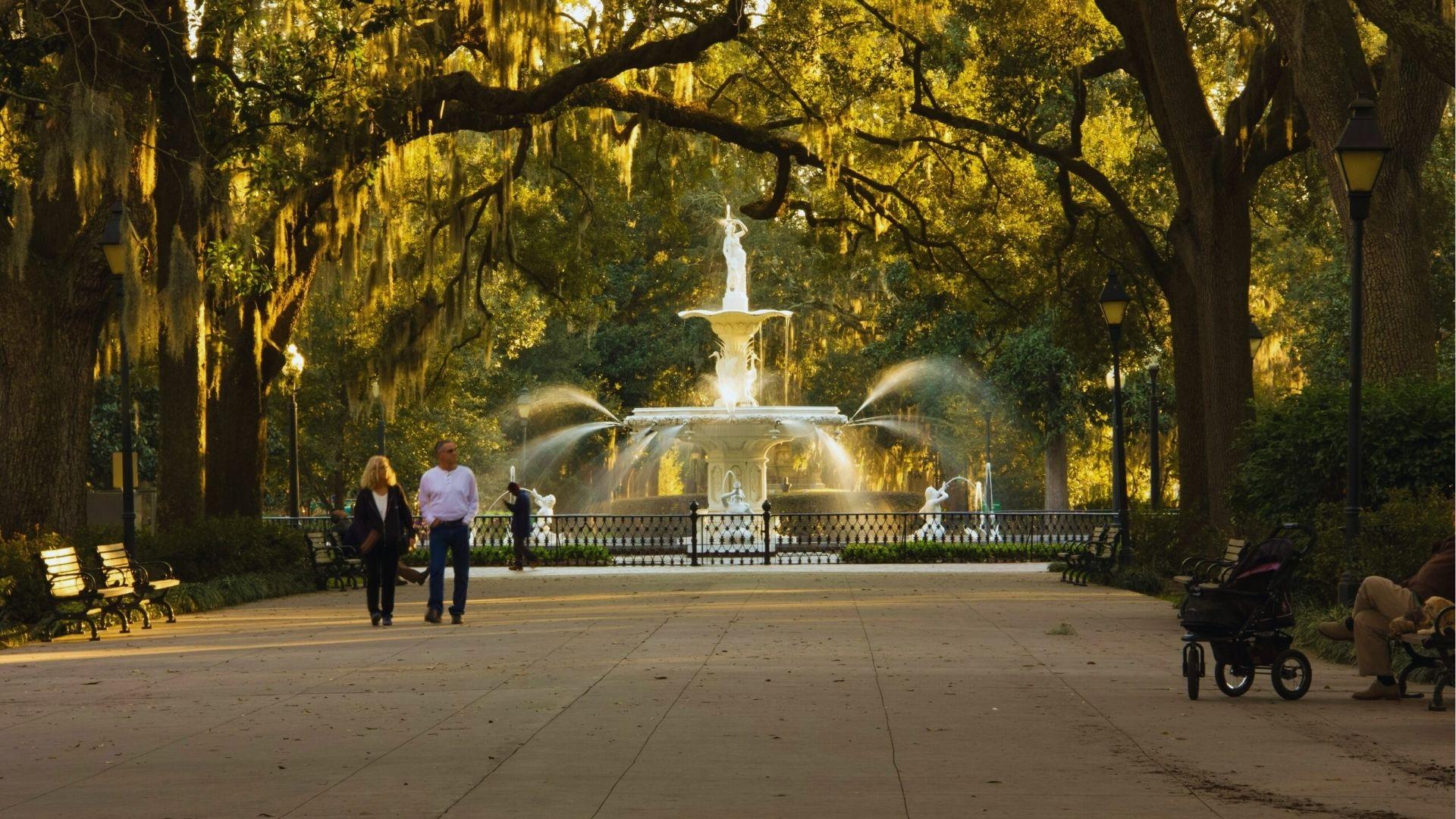 Forsyth Park fountain surrounded by moss-draped oaks with visitors strolling on a sunny afternoon pathway today.