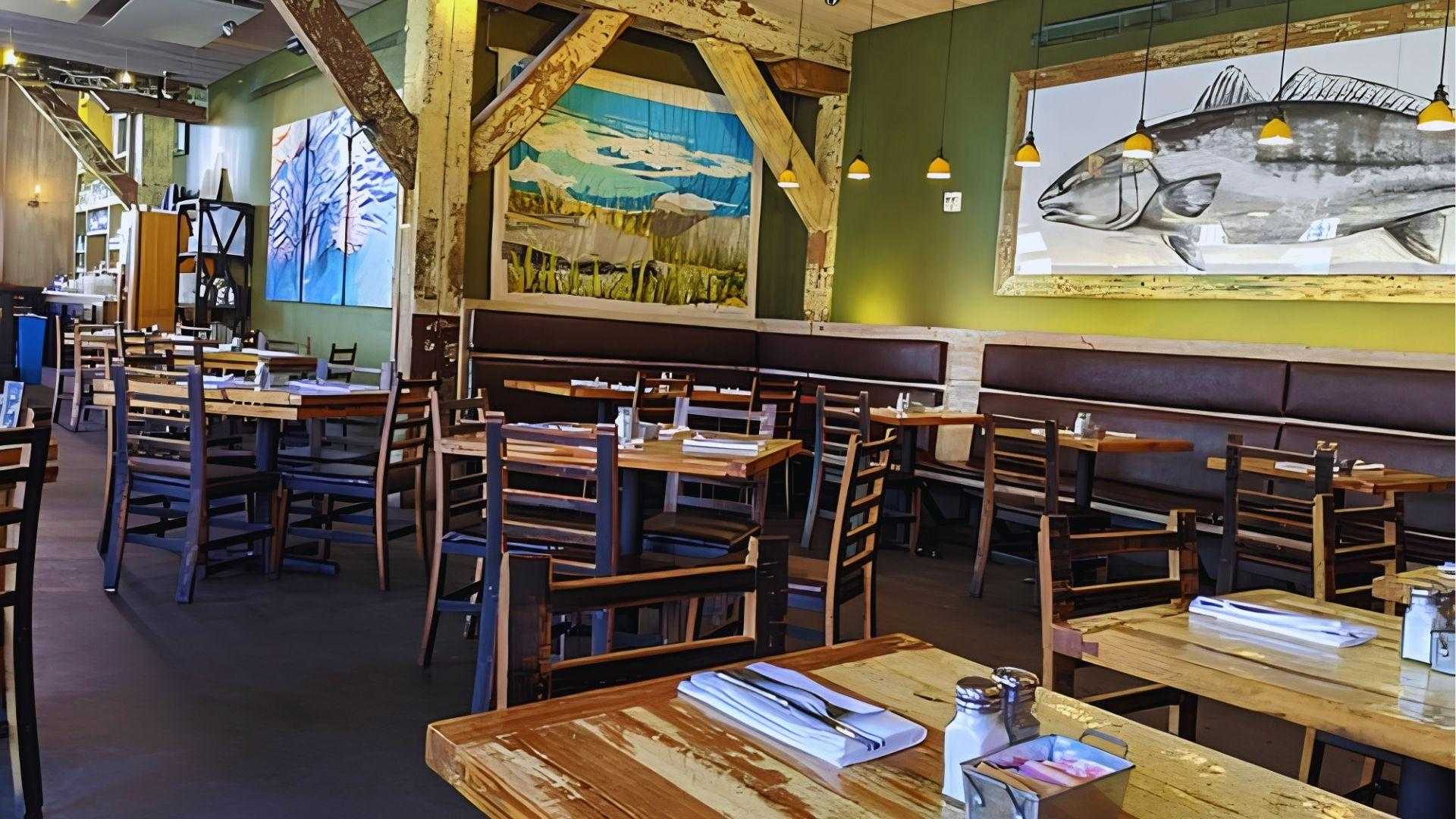 Seafood restaurant interior with wooden tables, dark chairs and large fish paintings on a green wall.