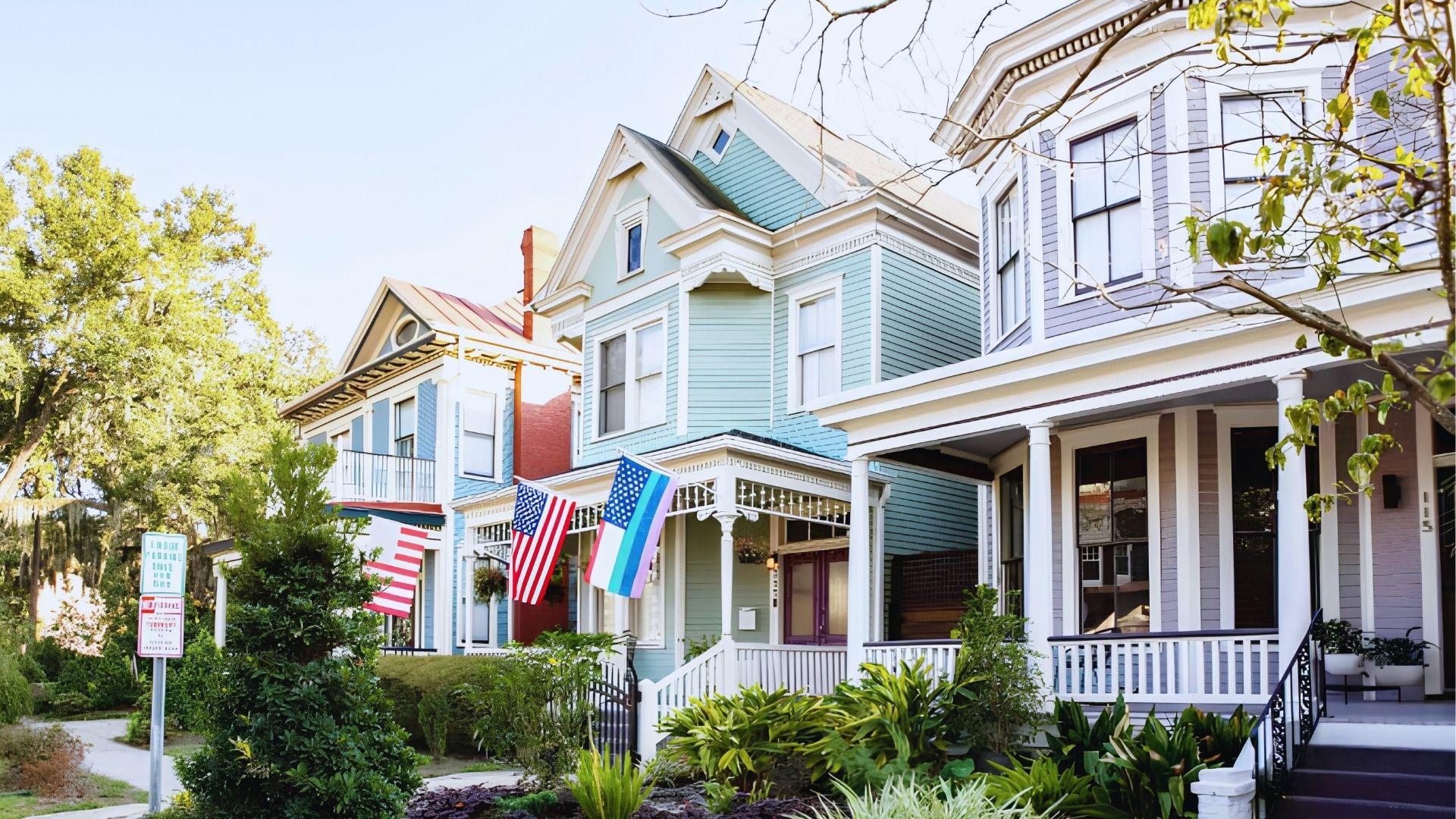 Pastel Victorian houses with American flags, lush greenery, charming porches on sunny Savannah street today.