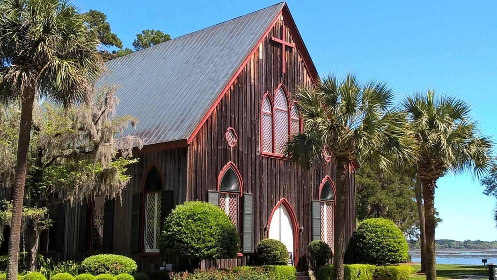 Historic dark wood chapel with red trim, pointed arches, cross on gable, palms and waterfront under blue sky