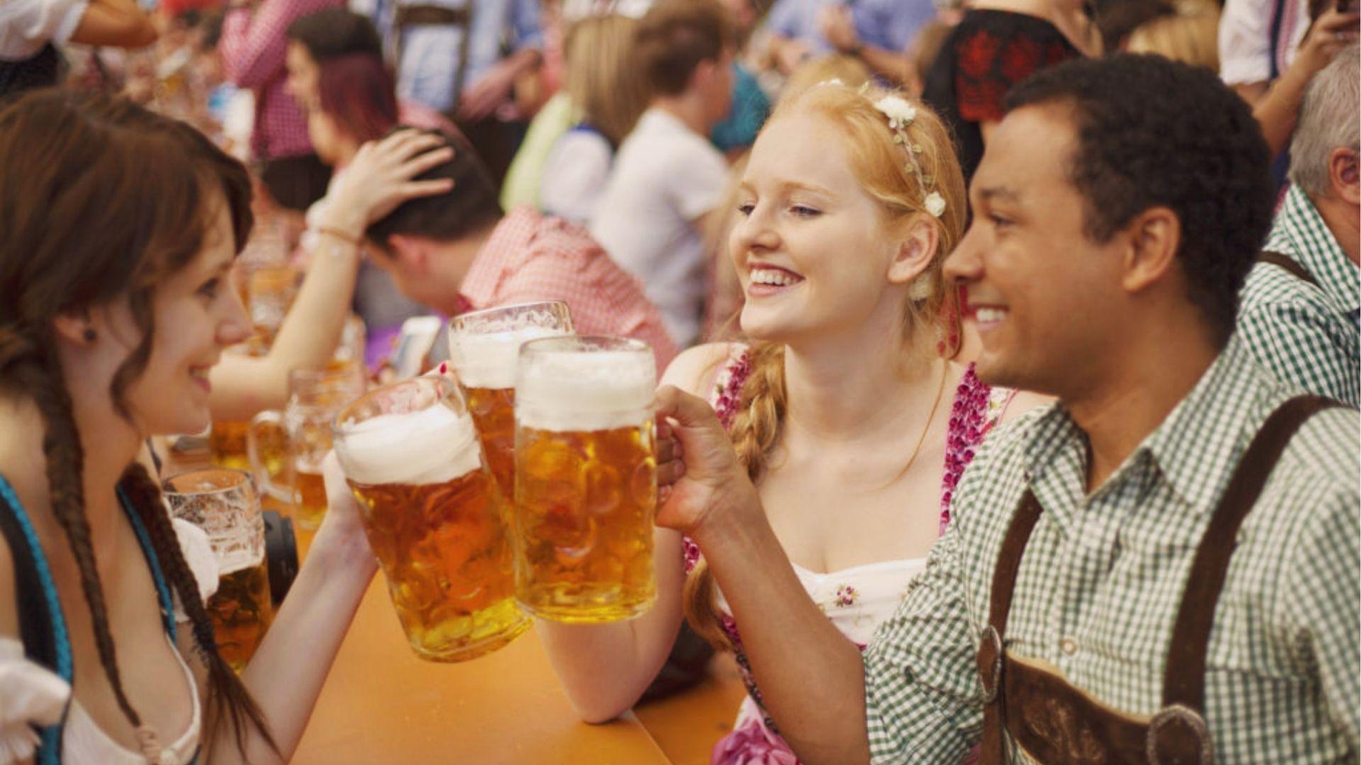 Friends in traditional Bavarian attire toasting large beer mugs at a crowded Oktoberfest beer garden table.