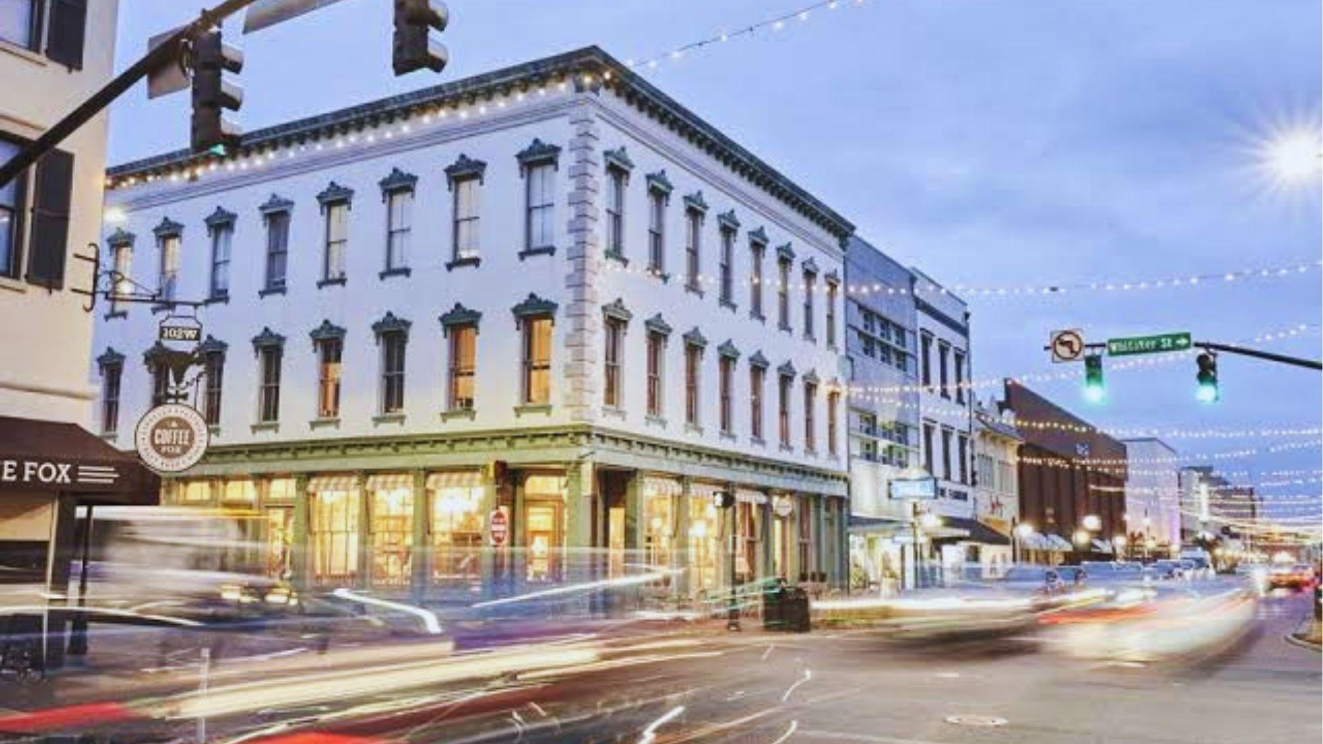 White historic corner building decorated with string lights at dusk with blurred traffic light trails.