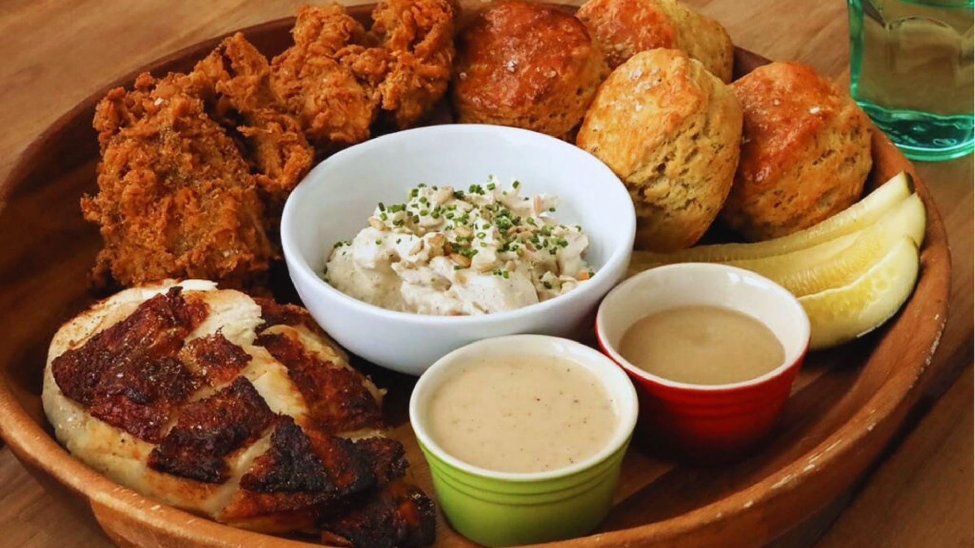 Wooden platter with crispy fried chicken biscuits creamy chicken salad pickles and two dipping sauces.