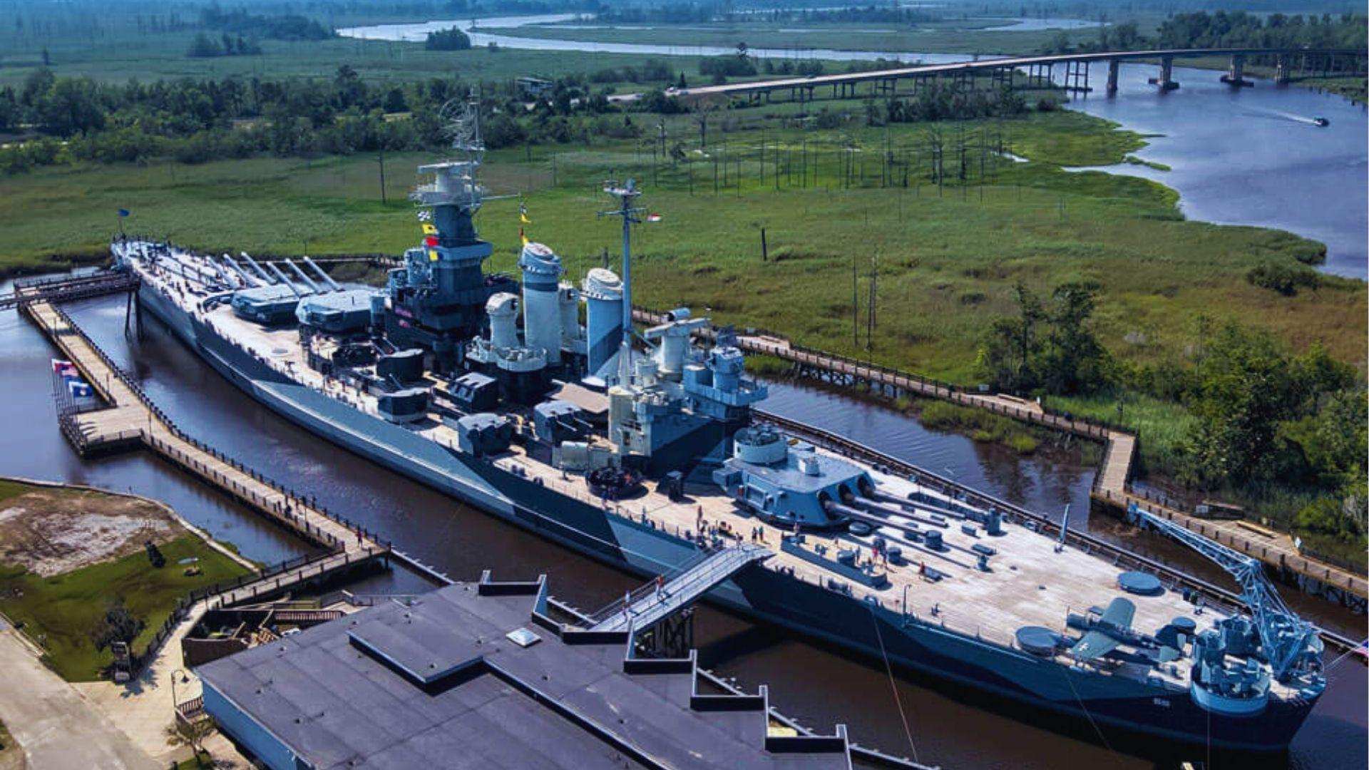 View of historic Battleship North Carolina docked in marshy river with wooden walkways and bridge.