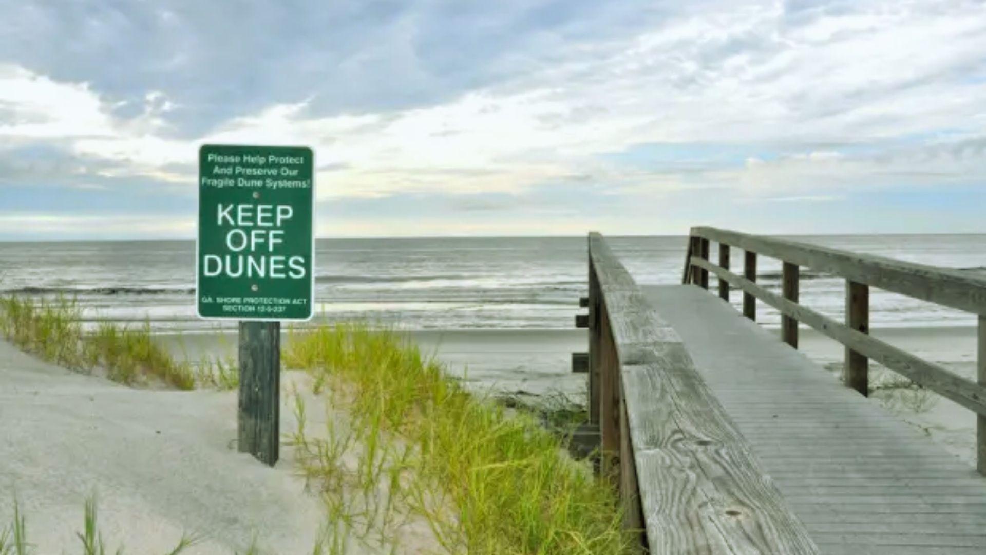 Wooden boardwalk leading to beach beside Keep Off Dunes sign and grassy sand dunes under cloudy sky