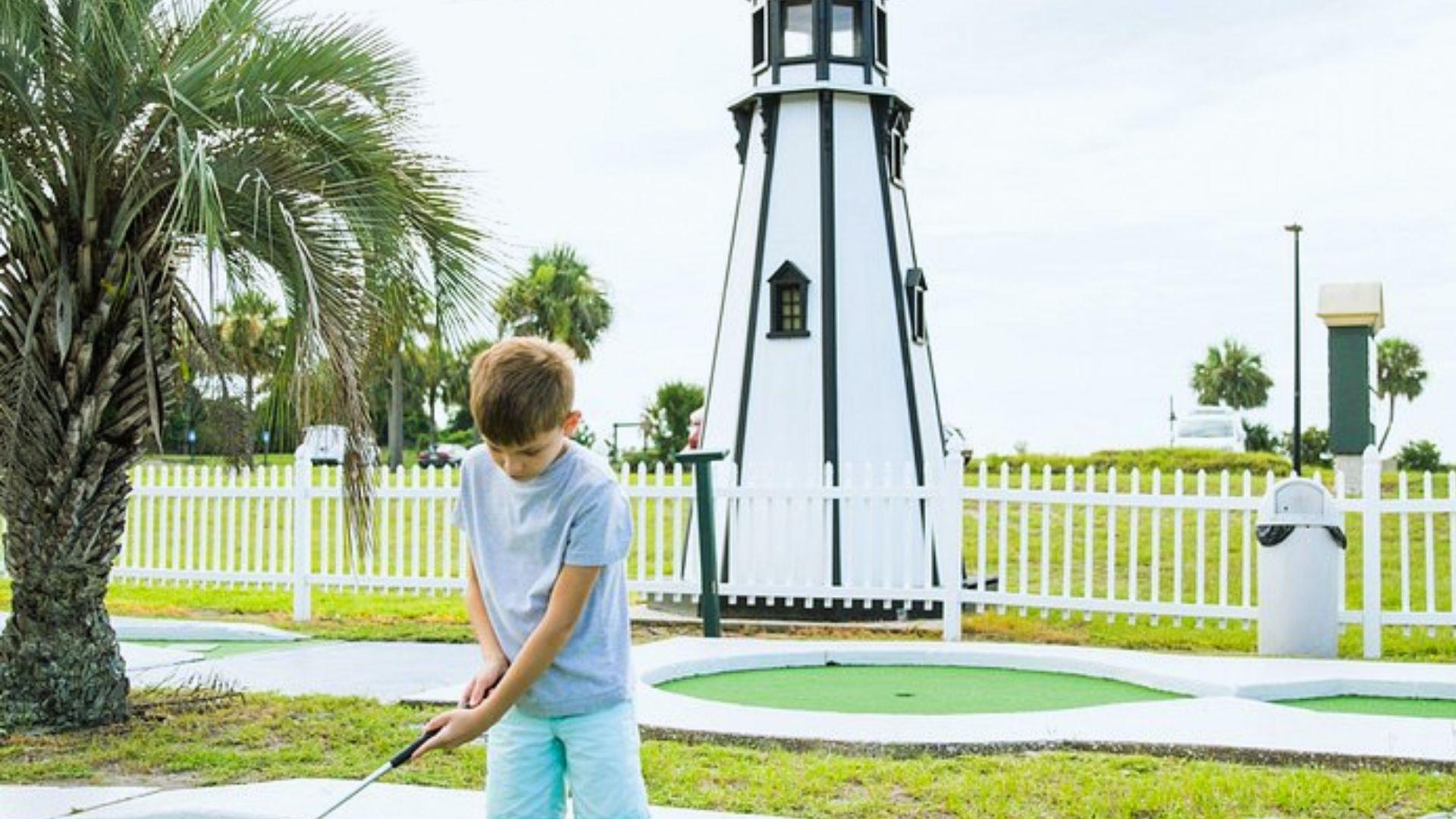 Young boy putting on mini golf course with lighthouse, palm tree and white fence behind