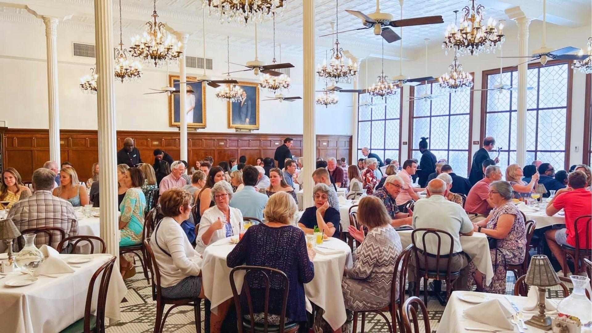 Elegant dining room with chandeliers portraits and guests seated at white-clothed tables enjoying meals together.
