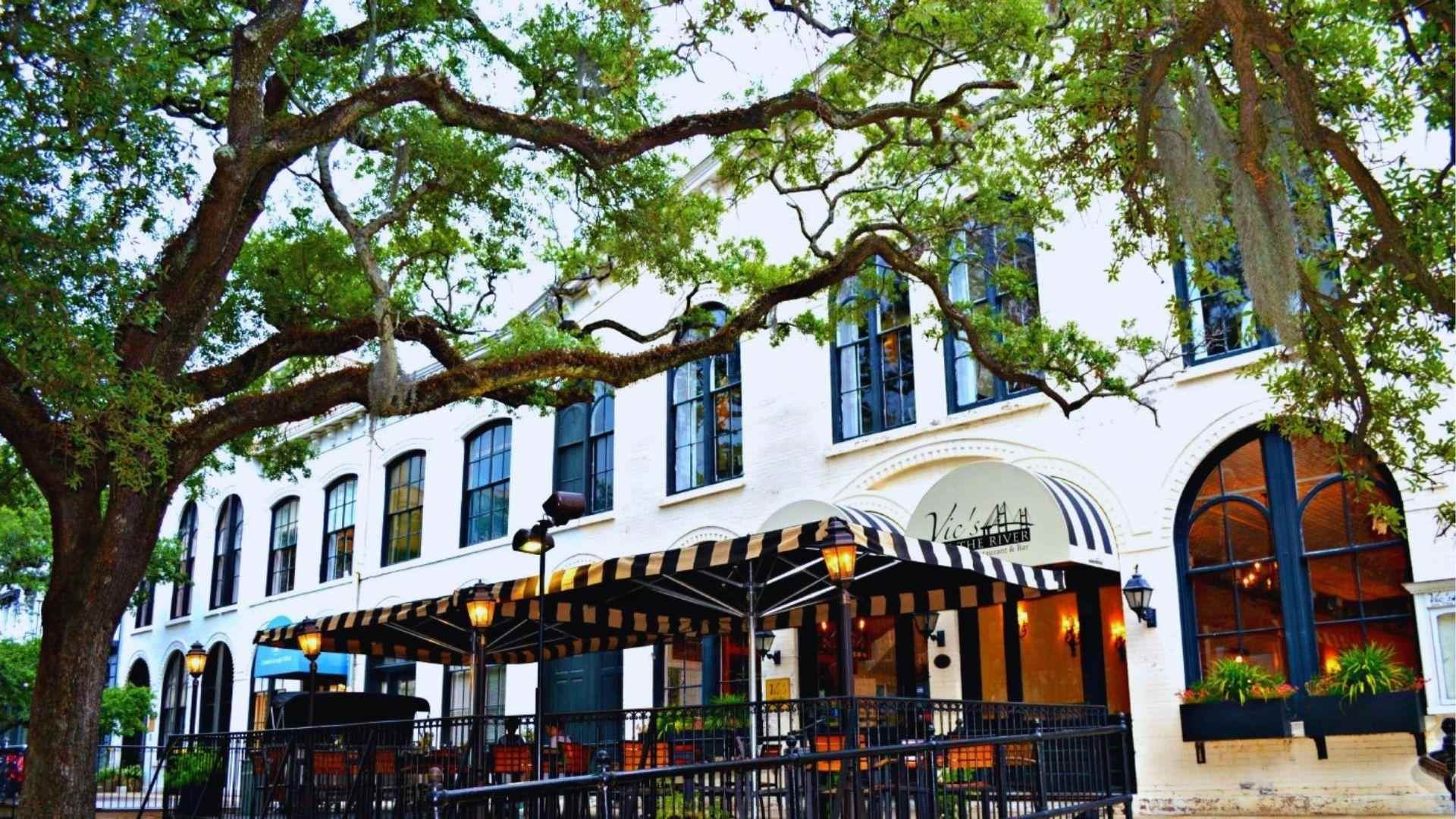 White historic Vic's on the River restaurant with striped awnings under large oak branches today.
