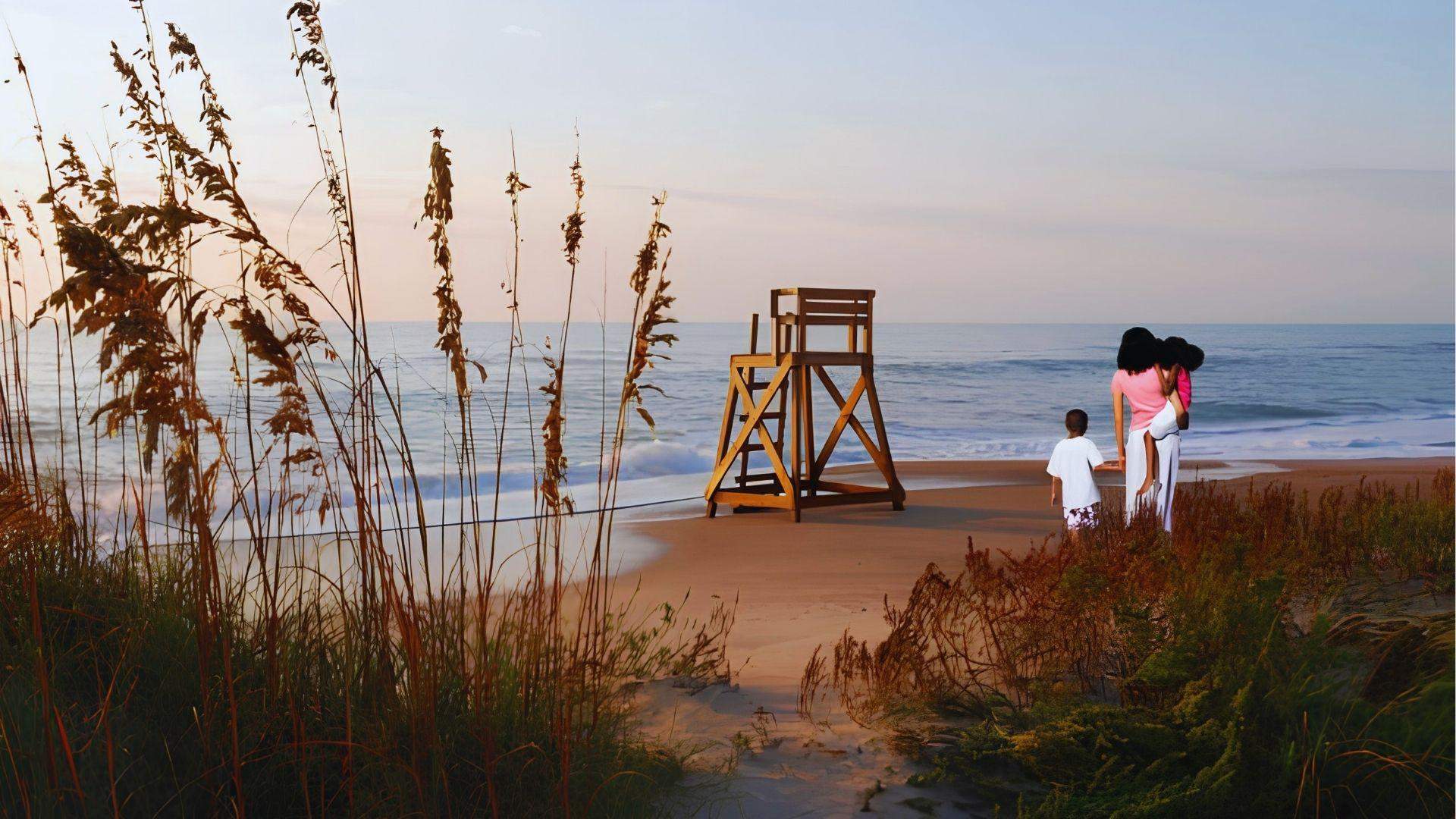 Family walking on beach at sunset near wooden lifeguard tower with sea oats in foreground.