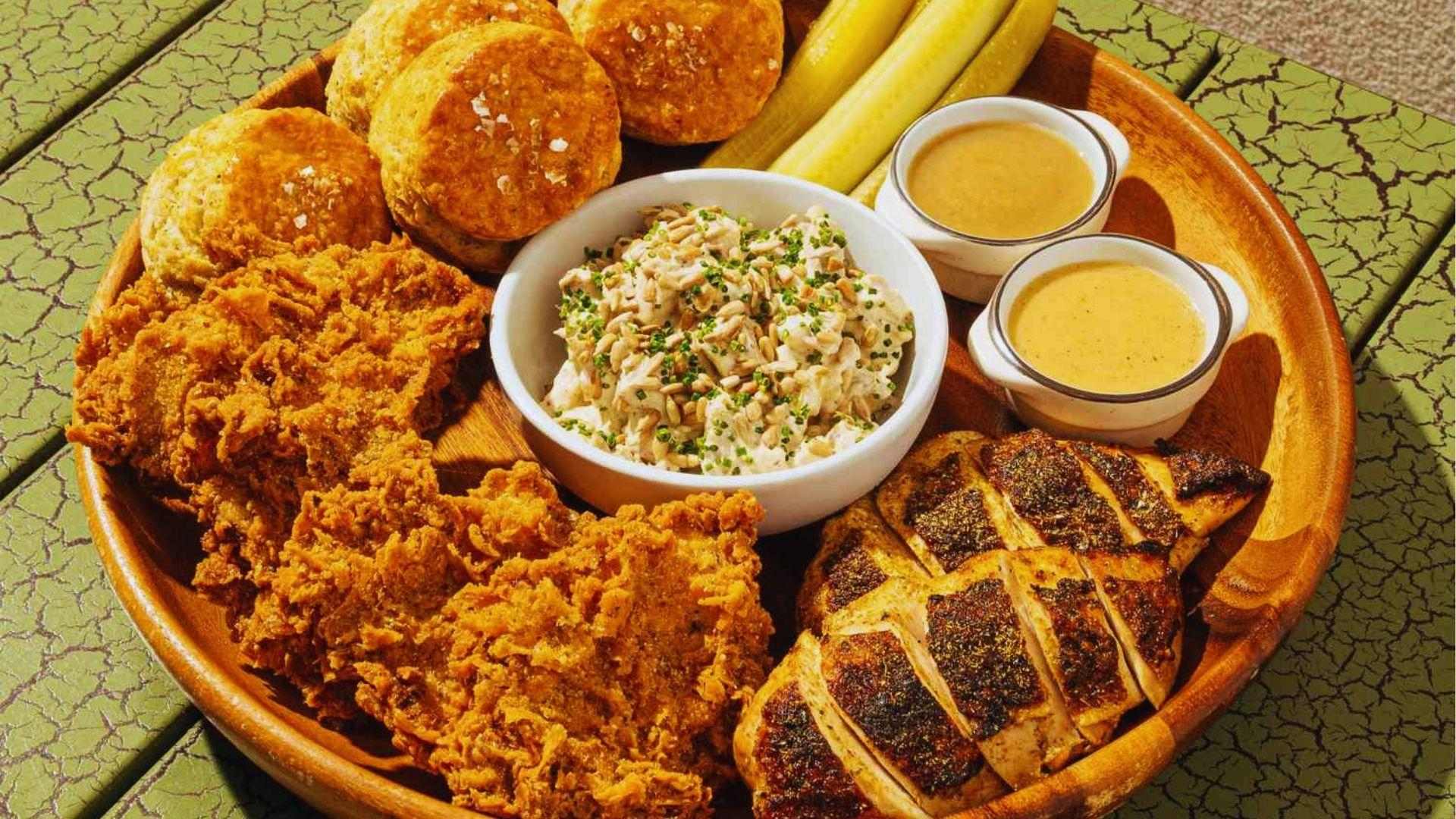 Wooden platter featuring crispy fried chicken grilled chicken biscuits coleslaw pickles and creamy dipping sauces.