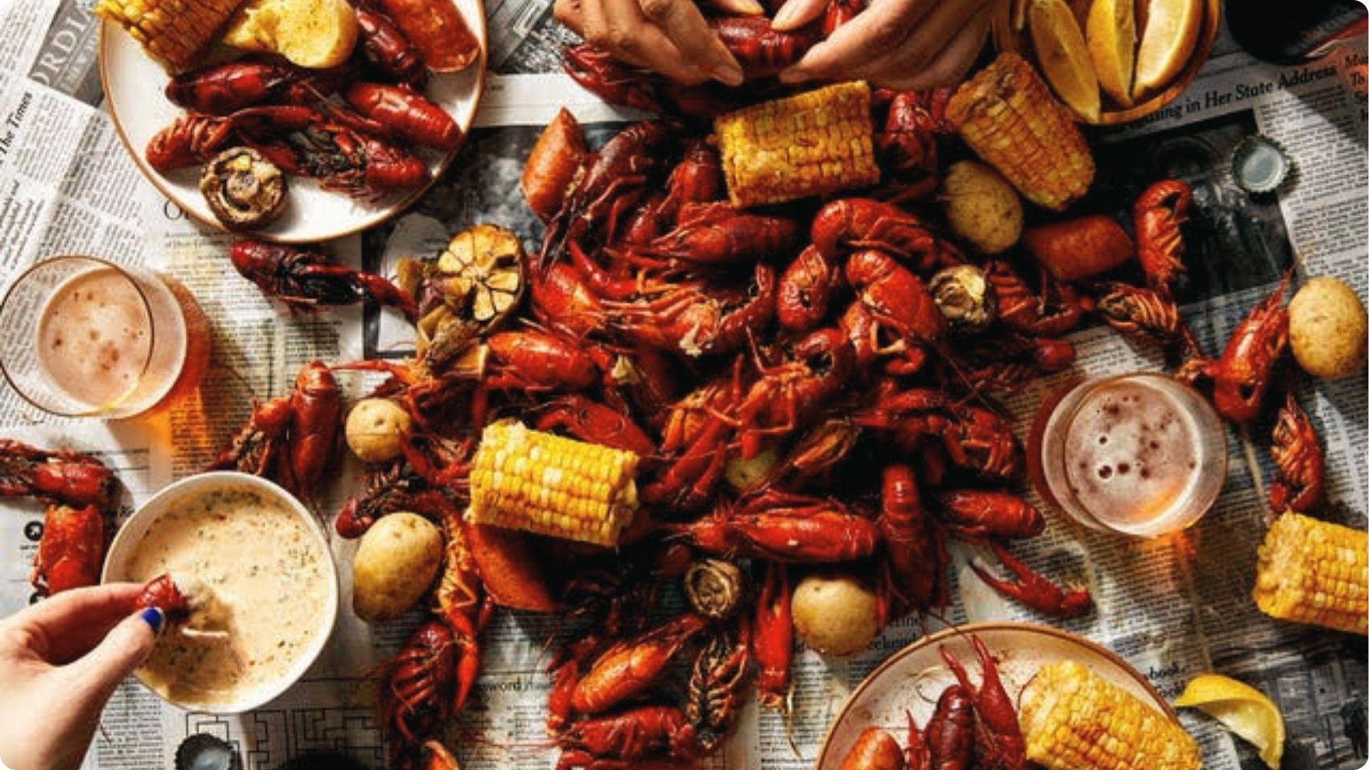 New Orleans crawfish boil spread with corn, potatoes, beer on newspaper, hands peeling spicy seafood.