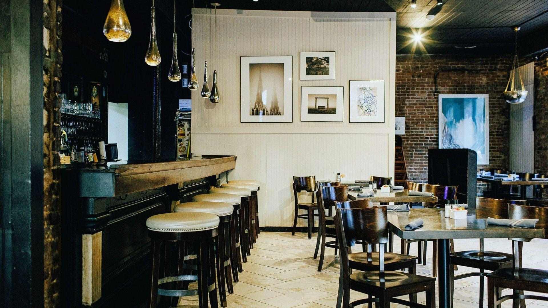 Modern restaurant bar with wooden stools, pendant lights, framed art, brick wall, and dining tables.